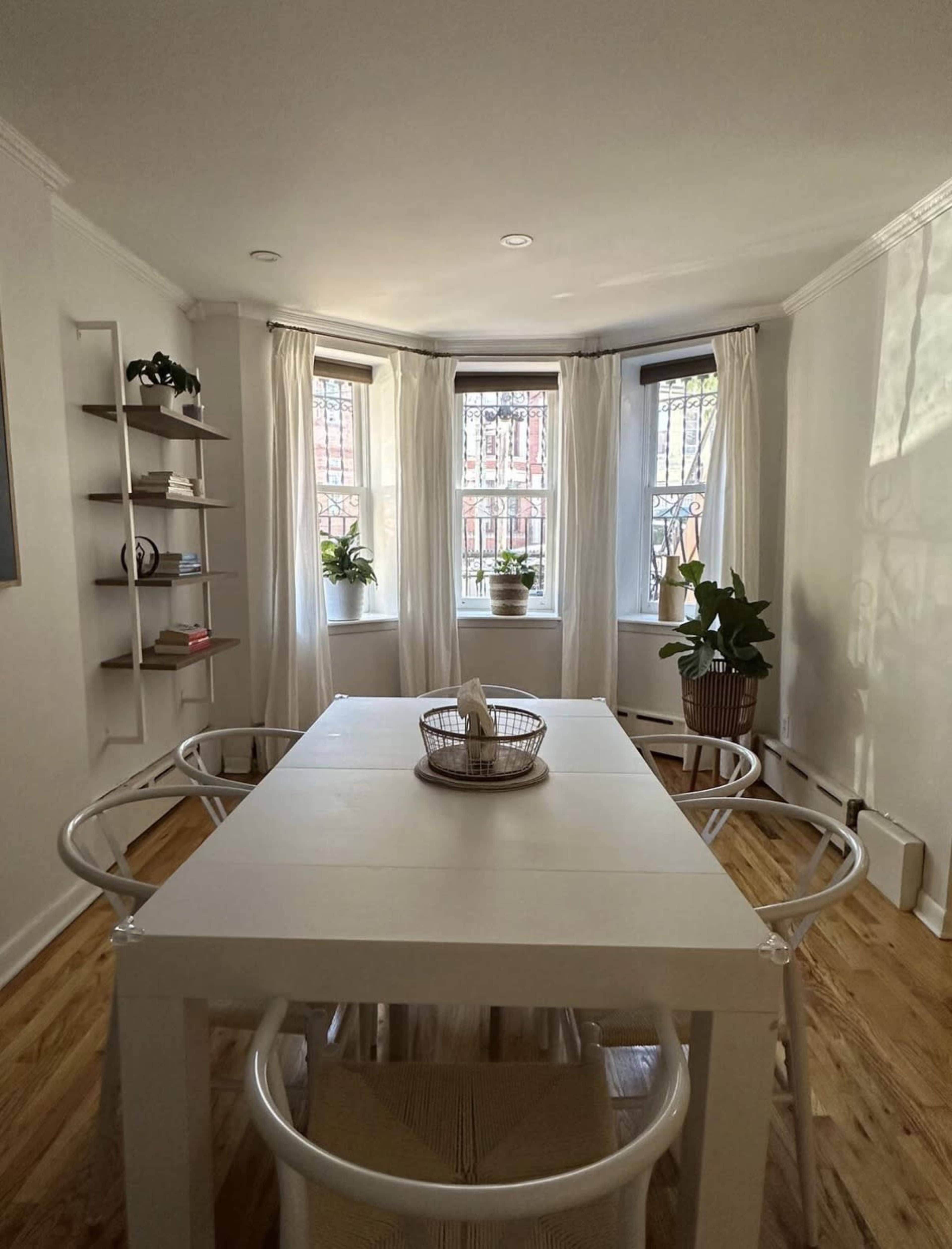 A bright dining area features a white table surrounded by chairs, with windows adorned by sheer curtains and plants near the glass.