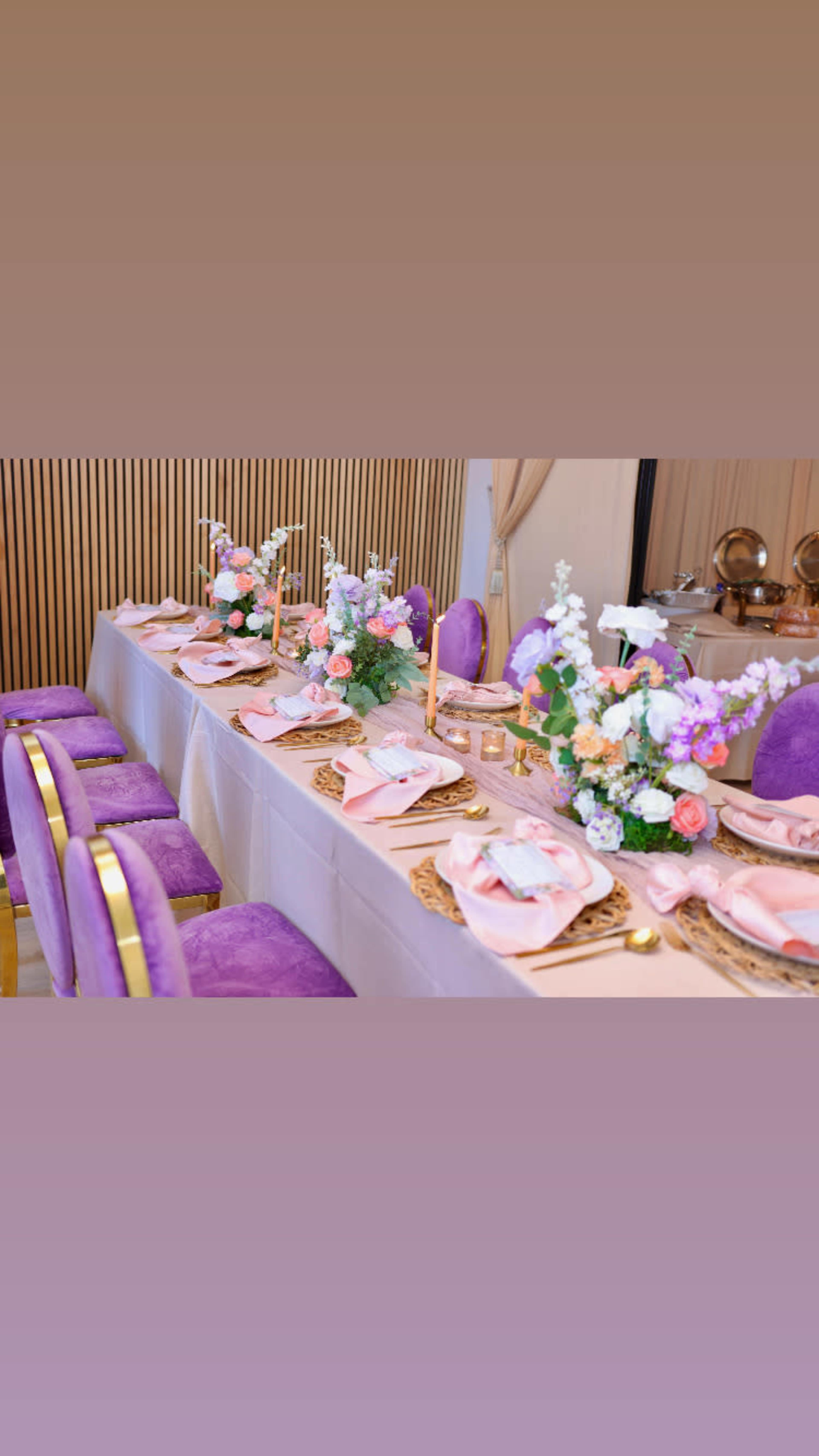 A long dining table is set with pink table linens, floral centerpieces, and purple chairs.