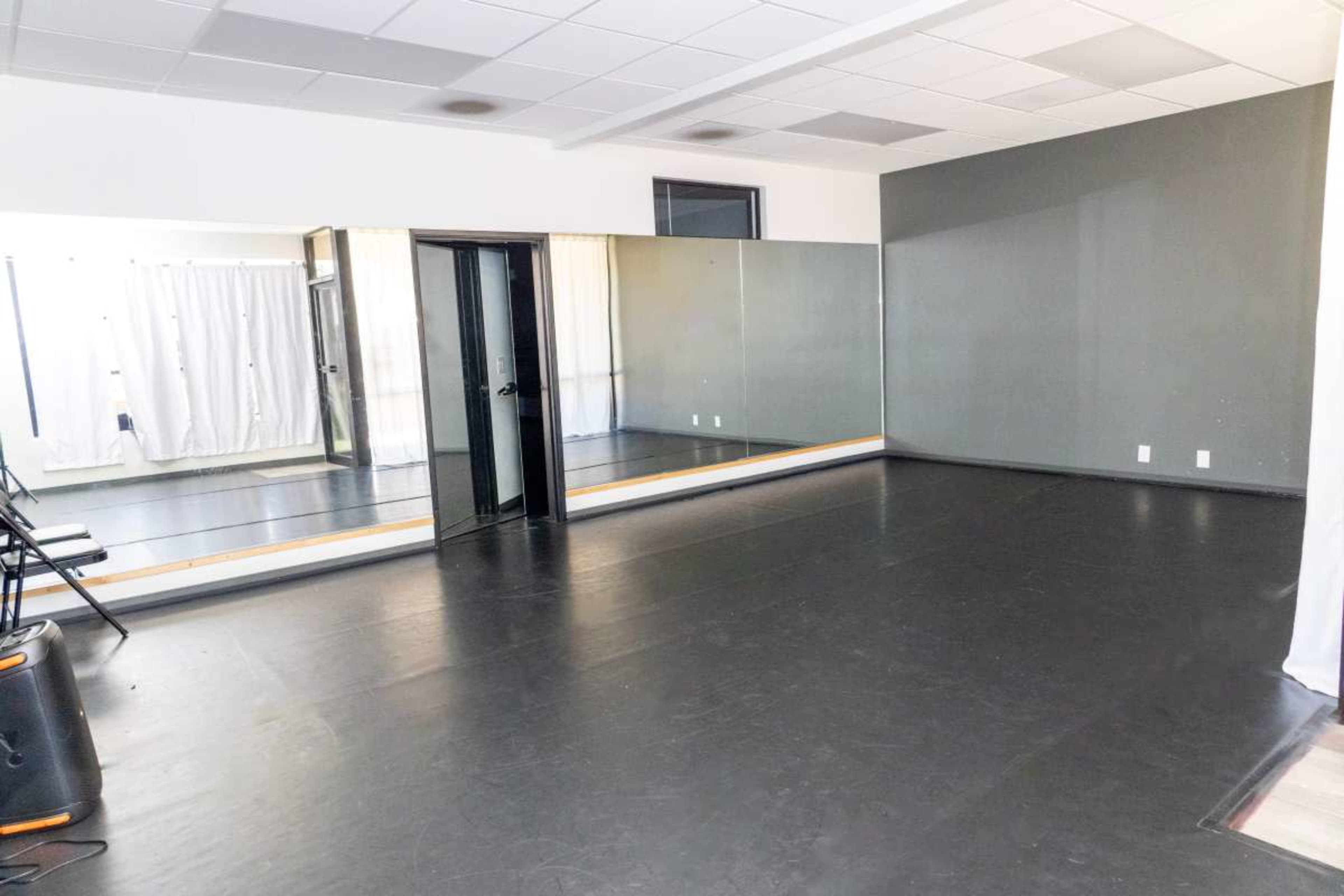The image shows a spacious dance studio with a black floor, large mirrors on one wall, and folding chairs arranged along the side.