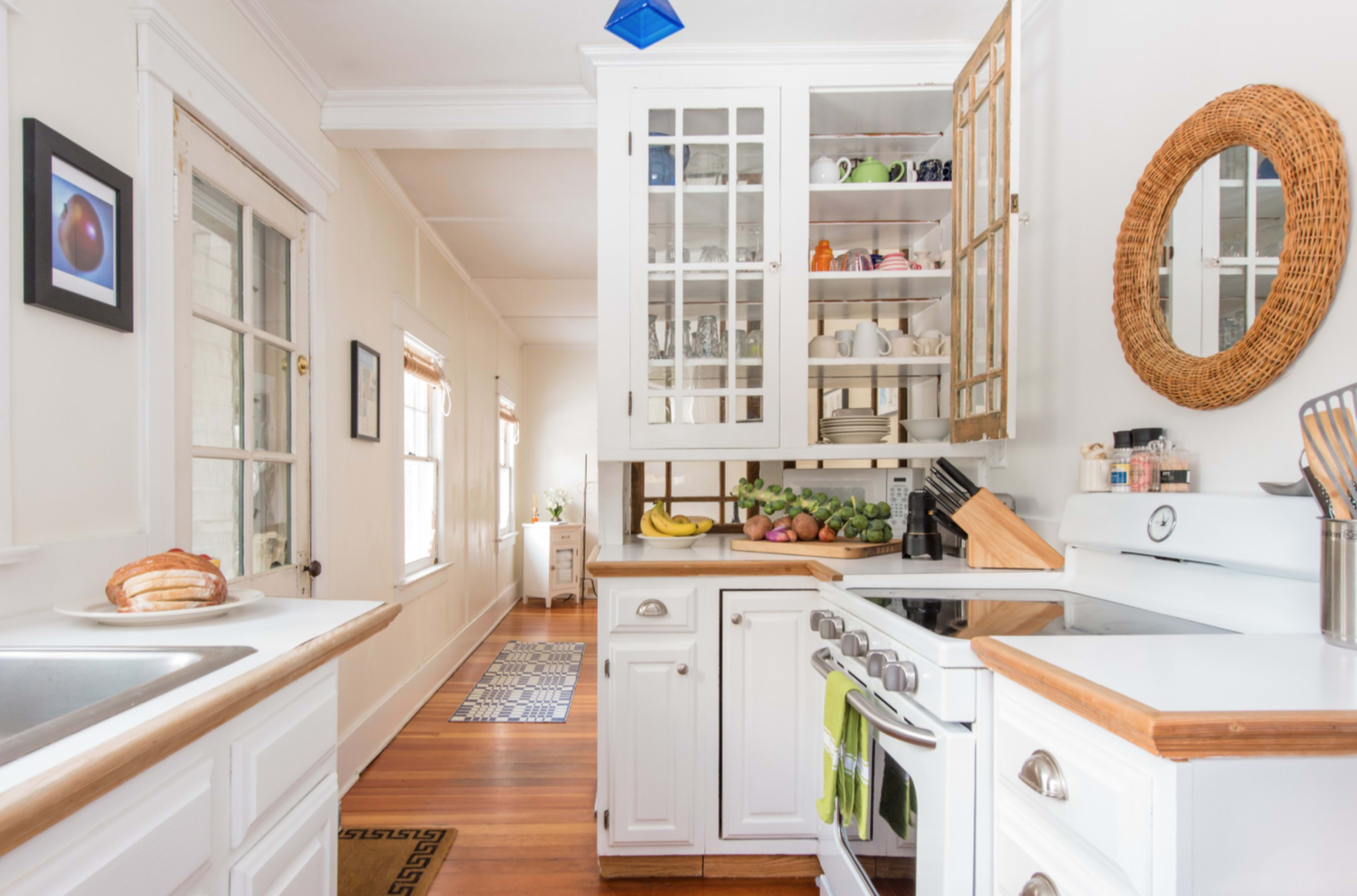 A bright kitchen features white cabinetry, a sink, a stove, and a glimpse of a hallway with a rug.