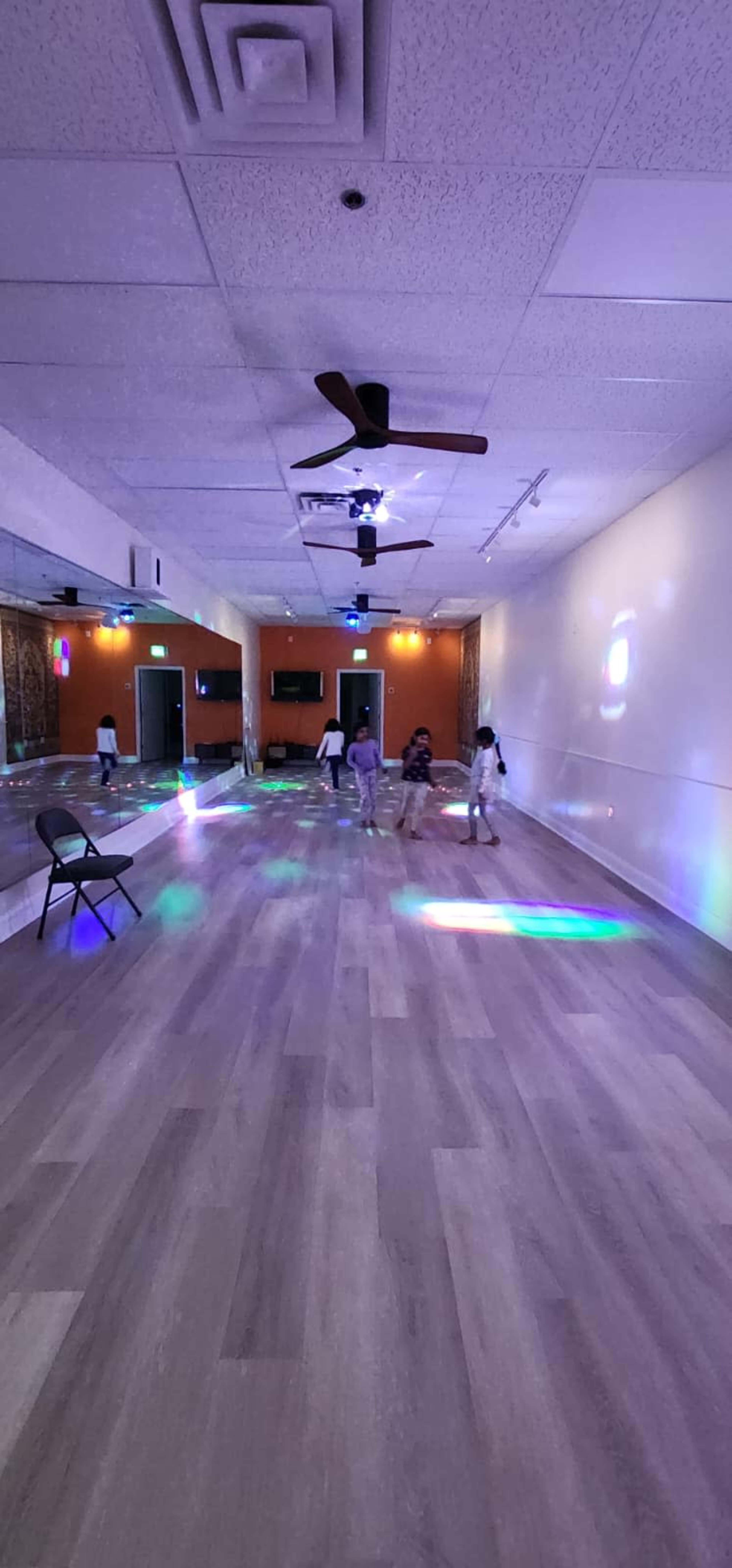 A spacious dance studio features a wooden floor, colorful lights projecting on the walls, and several children dancing in the center.