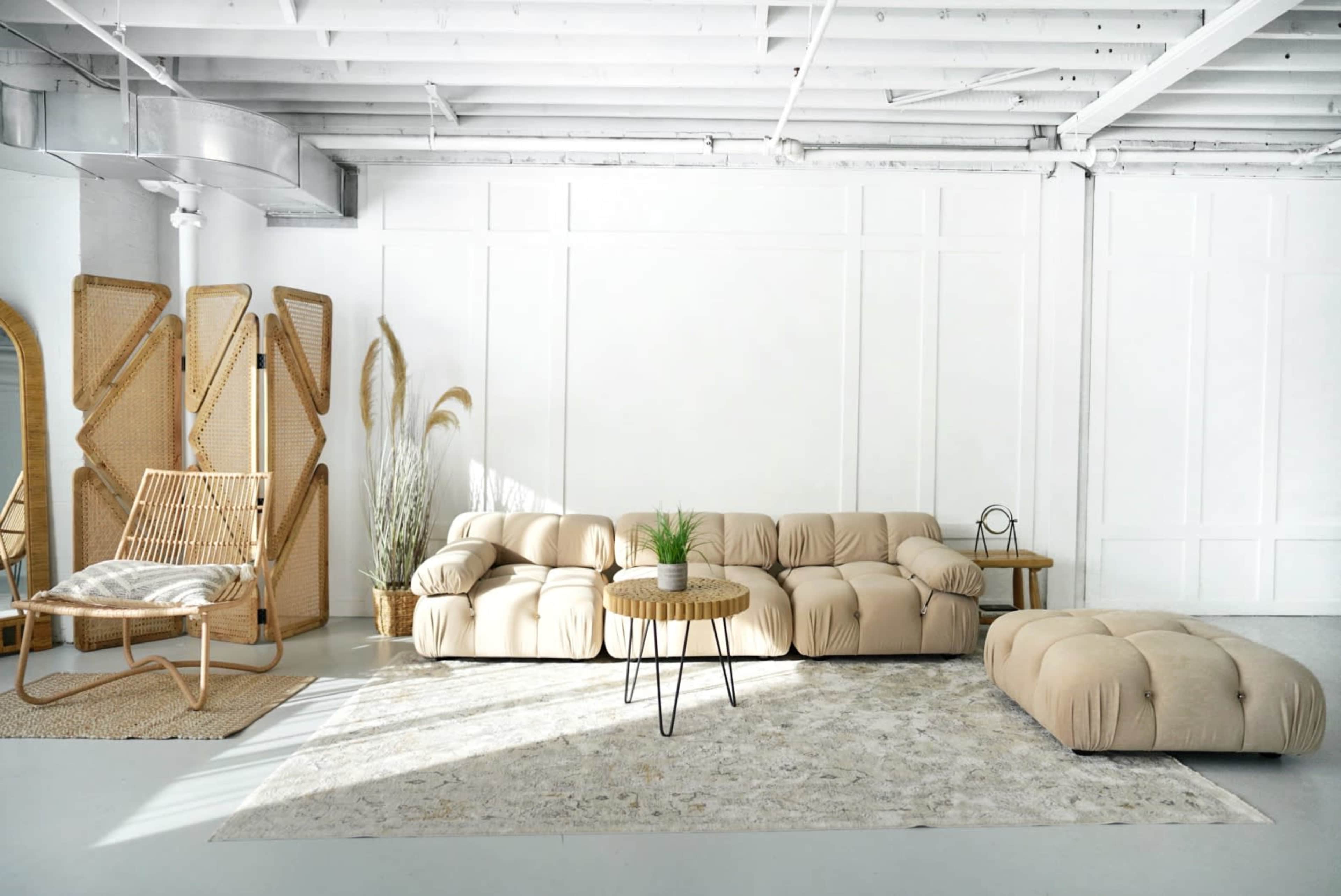 A spacious, light-filled room featuring a beige sectional sofa, a round wooden coffee table, a soft rug, and a rattan chair next to decorative plants.