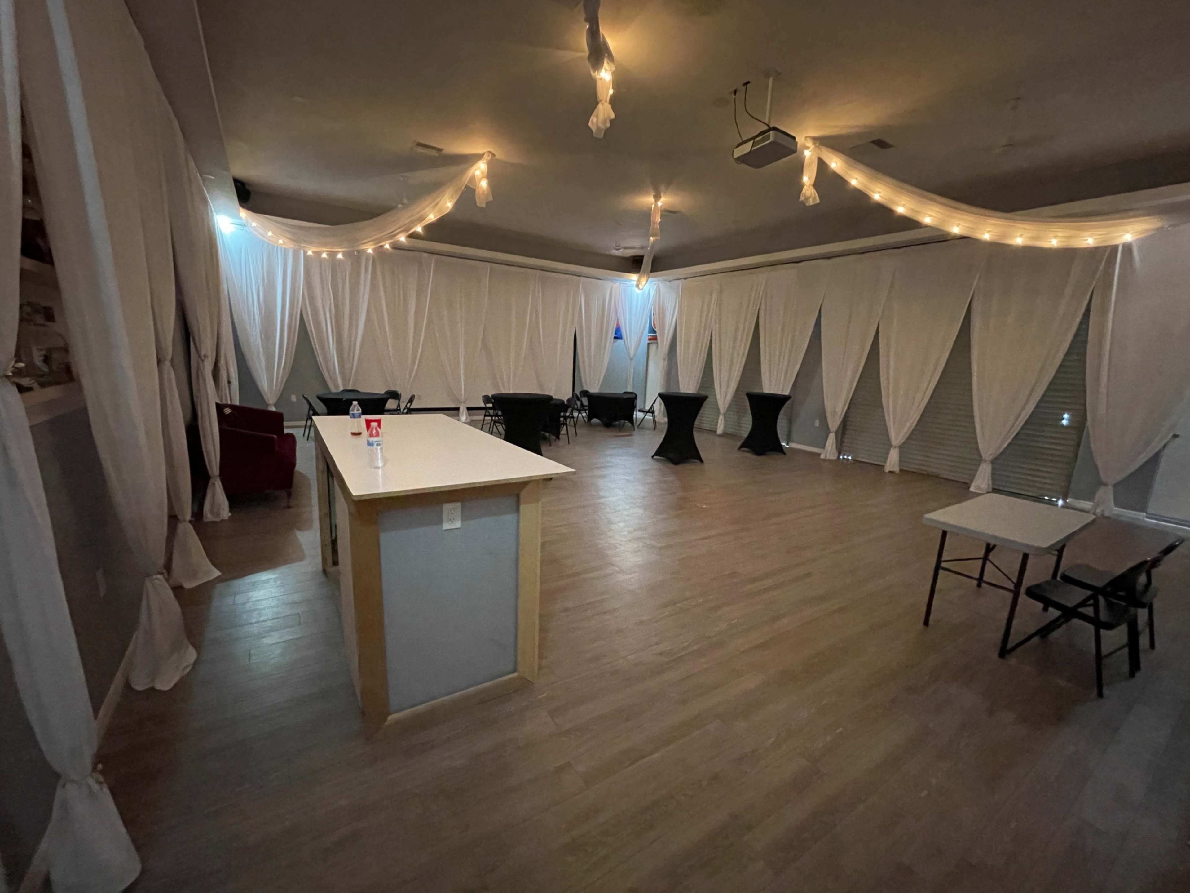 The image shows an indoor event space with draped white fabric, wooden flooring, a central bar area, and various tables arranged throughout.