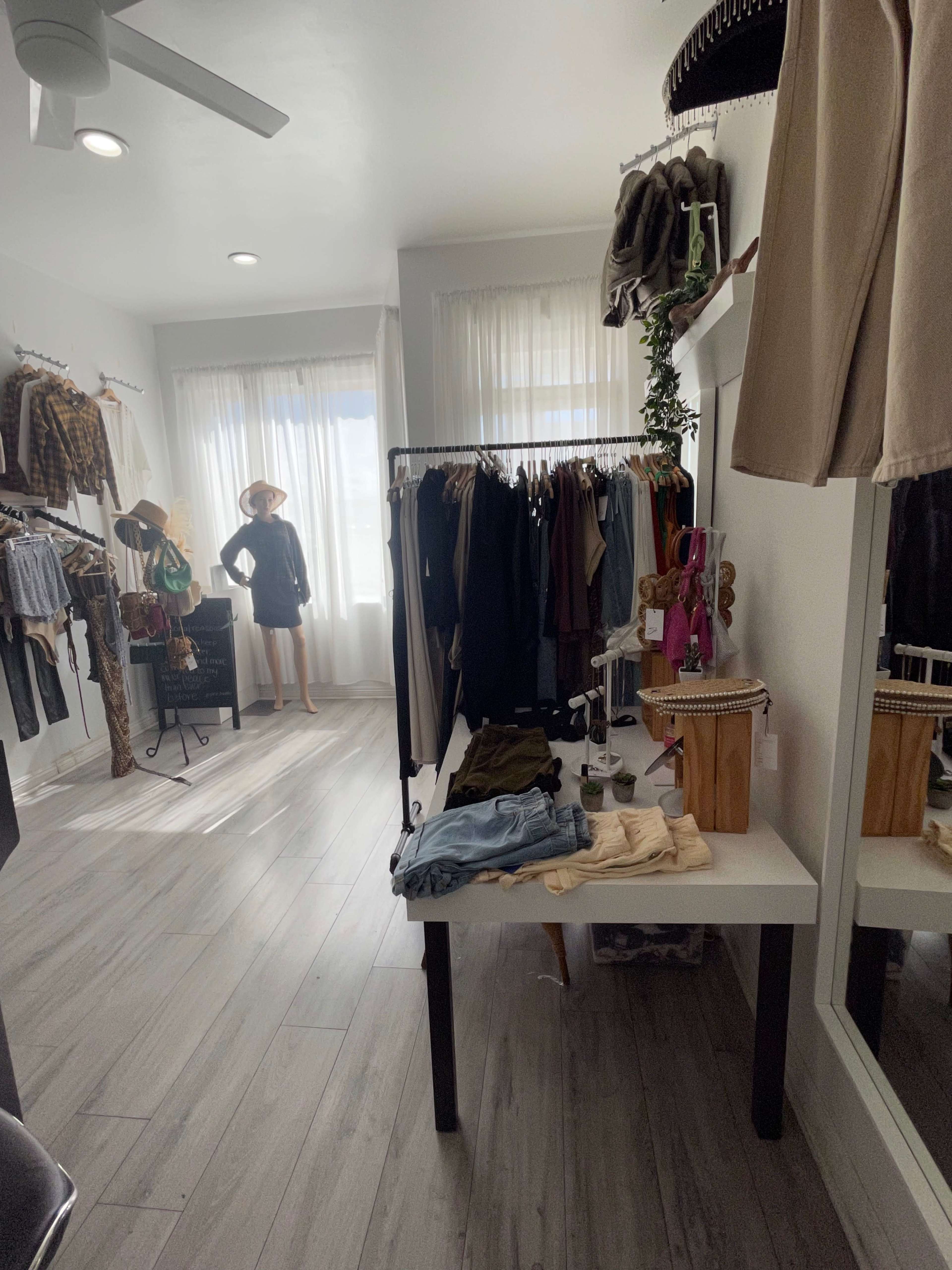 A boutique interior with clothing racks on the walls, a display table with folded garments, and a mannequin dressed in casual clothing near a window.