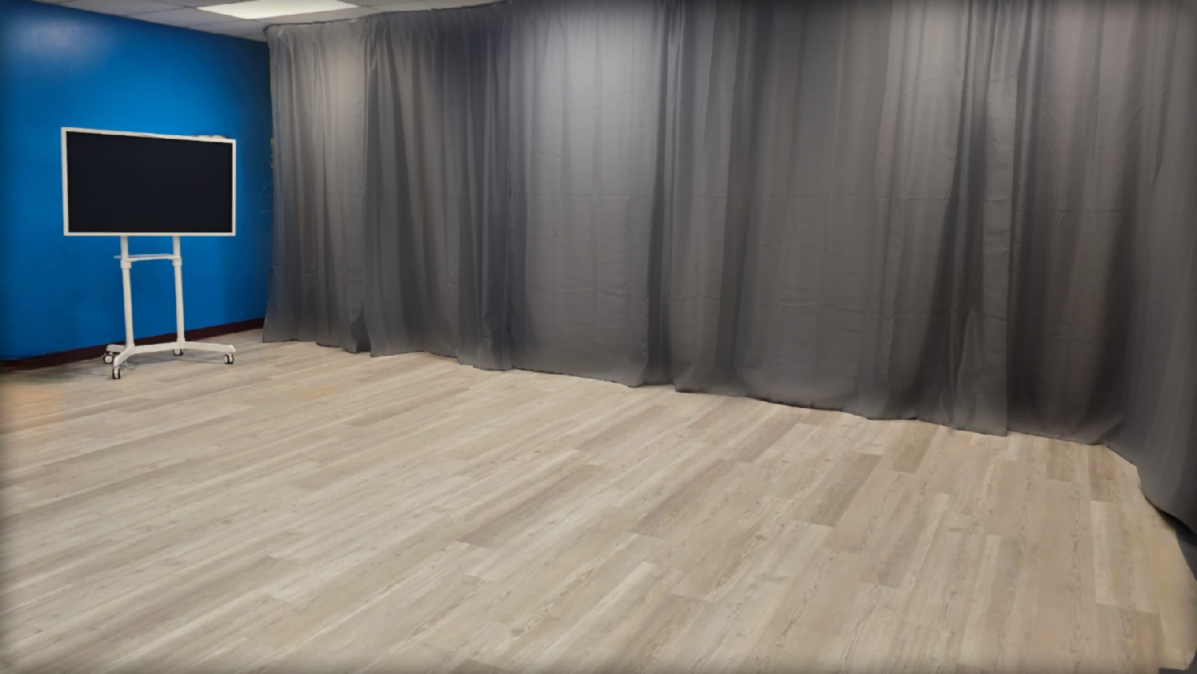 The image shows a room with grey curtains draping on one side and a large screen on a stand set against a blue wall.