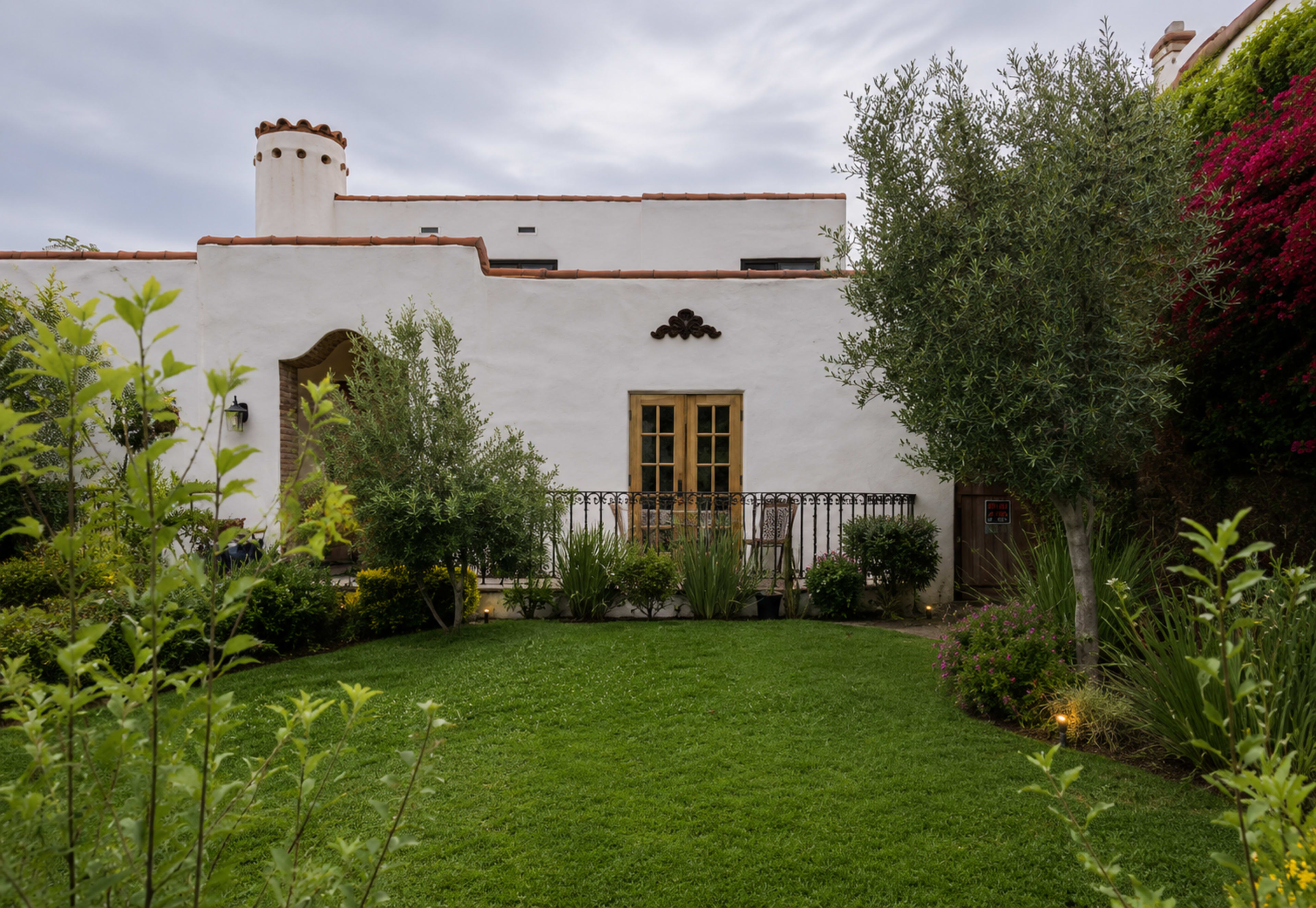 The image shows a white stucco house with a terracotta roof, featuring a balcony and surrounded by a well-maintained garden with green grass and various plants.