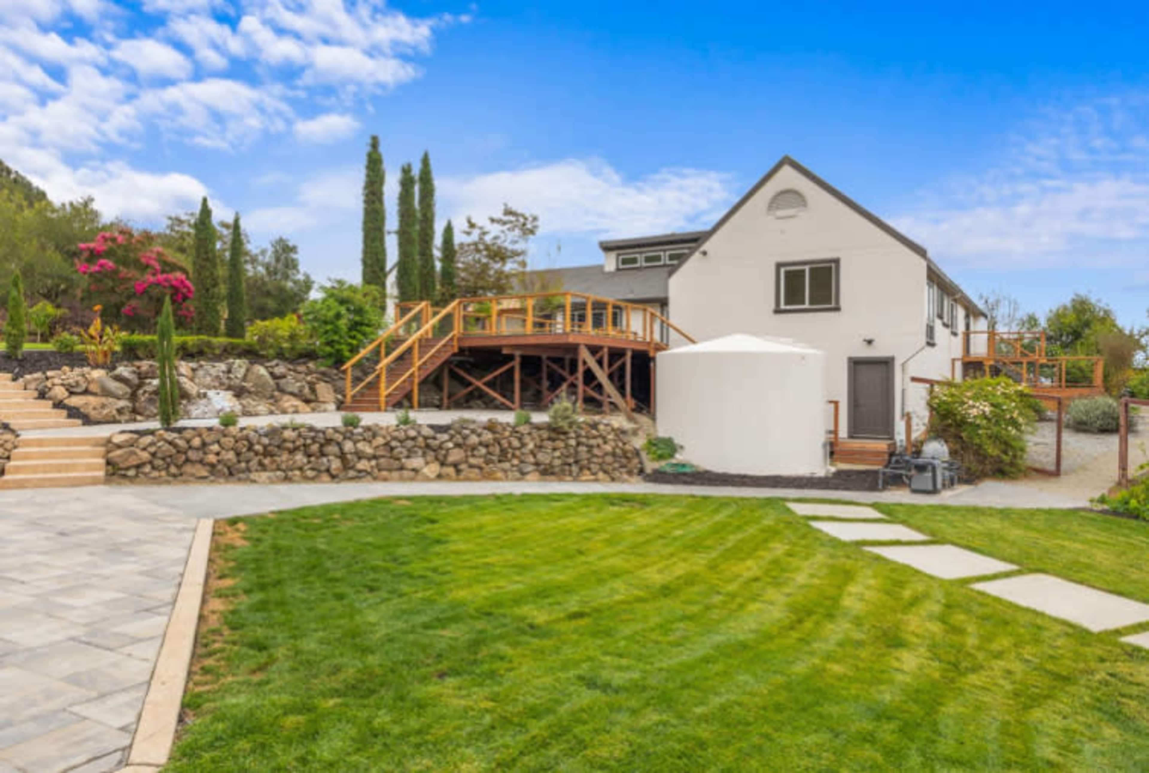 The image shows a large house with a wooden deck, surrounded by landscaped greenery and stone pathways.