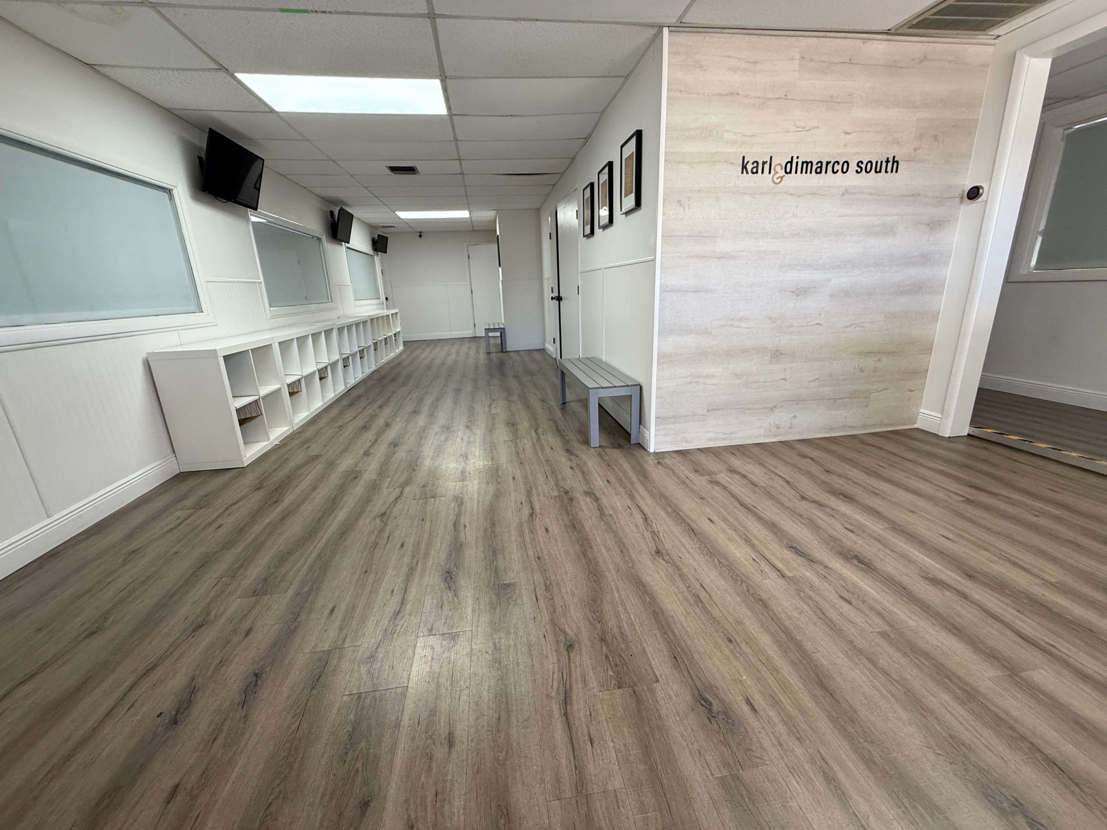 The image shows a spacious, well-lit corridor with wooden flooring, flanked by shelves on one side and a wall with framed pictures on the other.