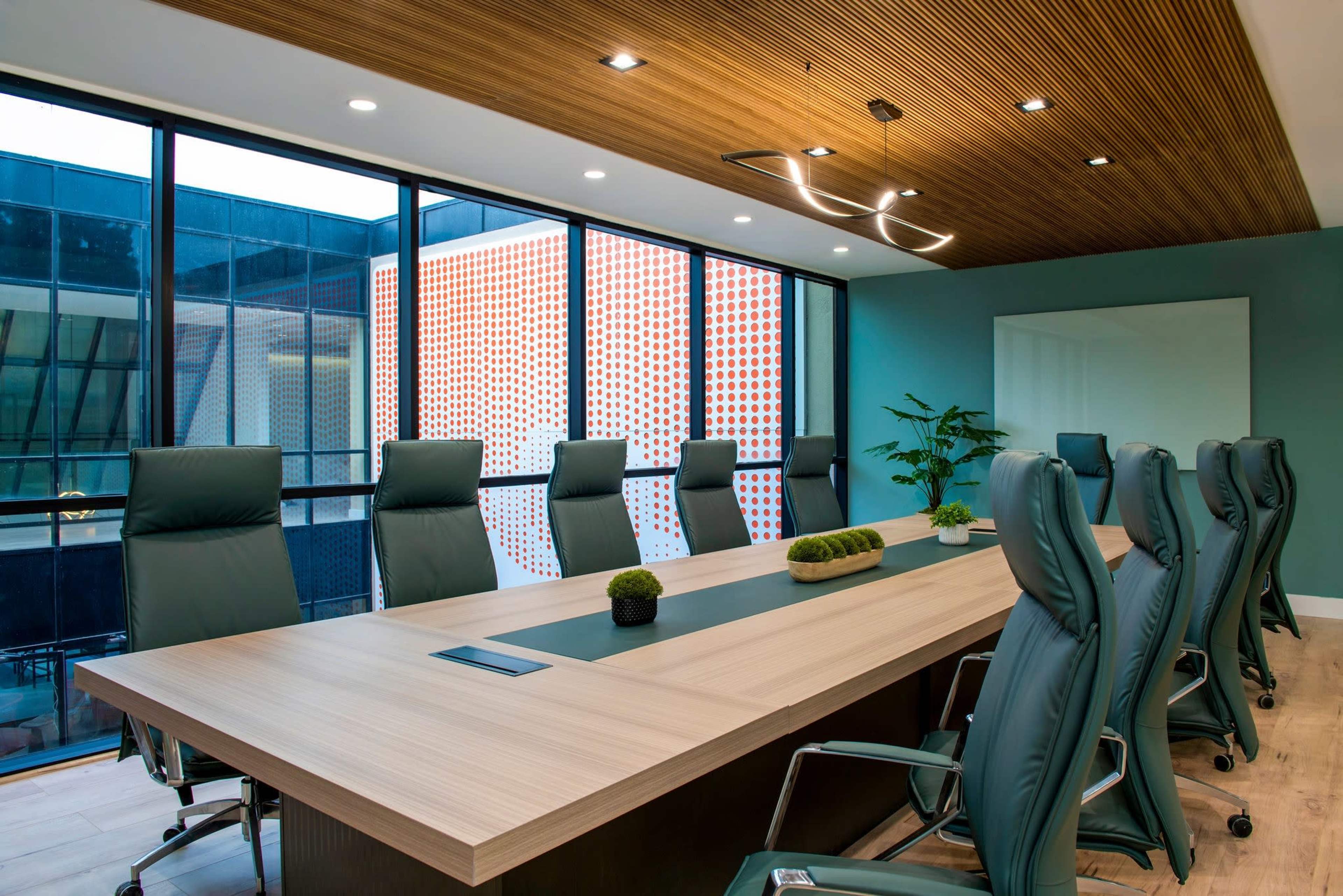 A conference room features a long wooden table surrounded by green ergonomic chairs, with large windows allowing natural light to enter.
