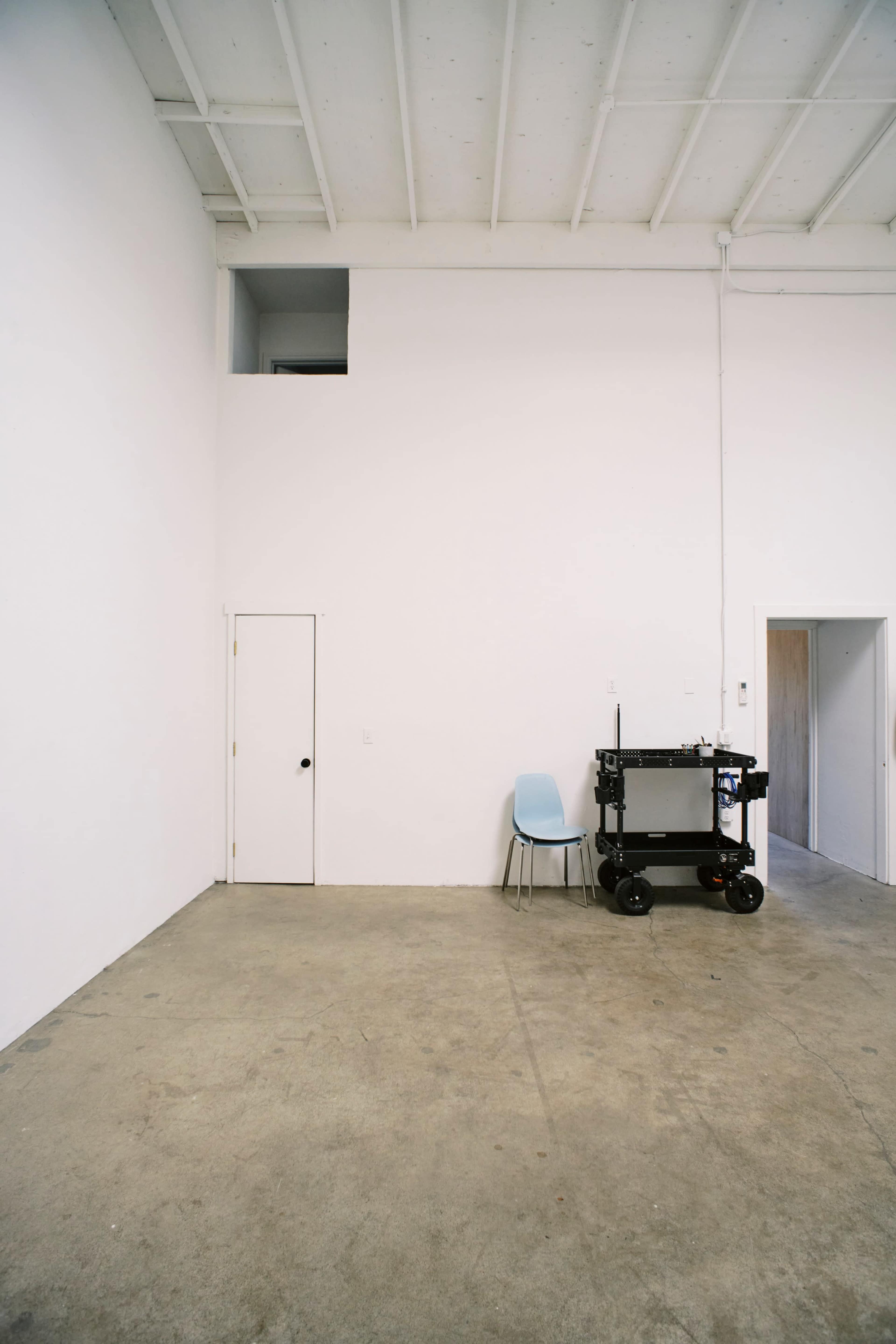 The image shows a spacious, empty room with white walls, a concrete floor, a light blue chair, and a cart positioned against one wall.