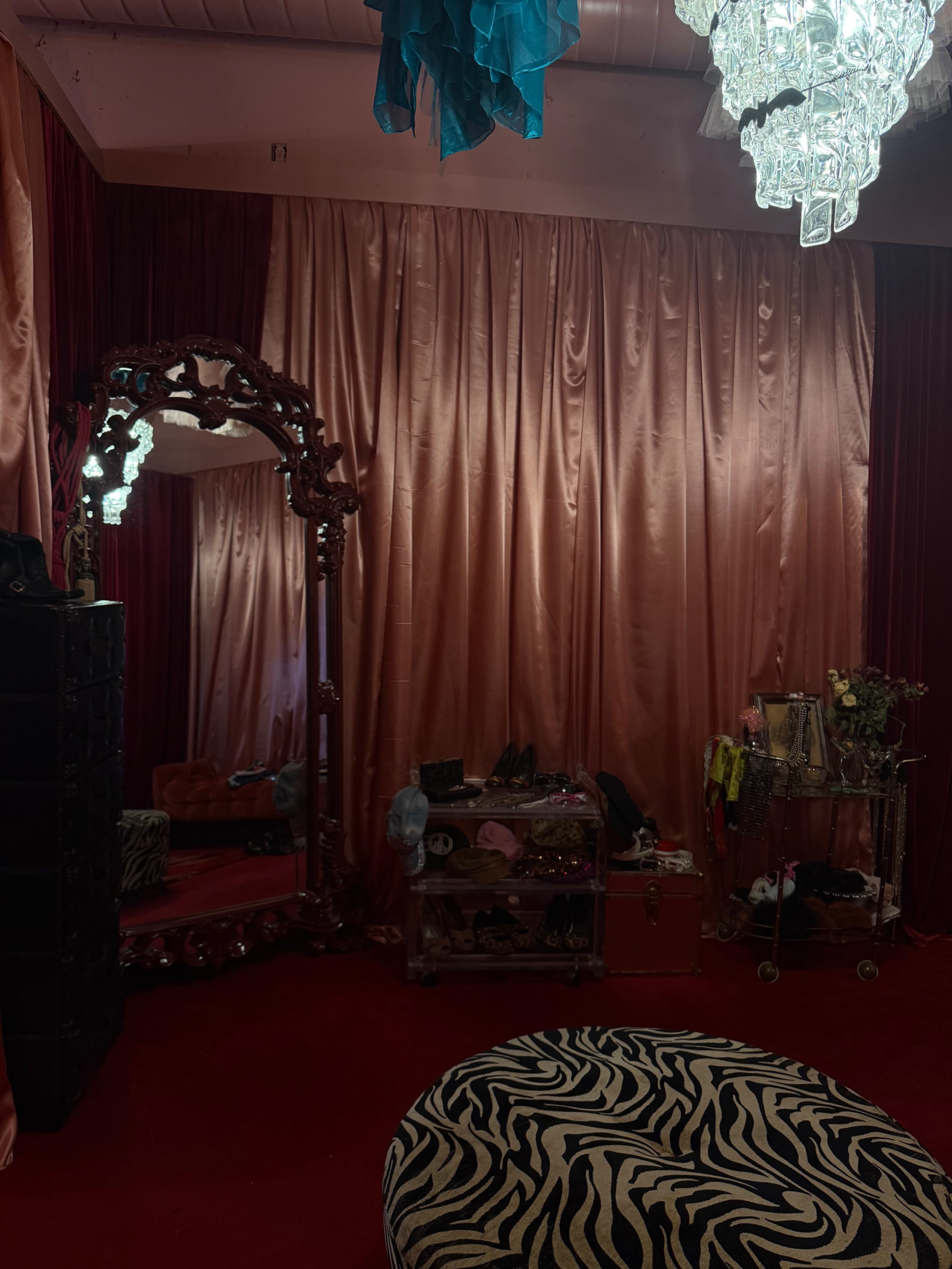 The room features a large ornate mirror, a chandelier, and walls draped in pink and red fabric, with a zebra-print stool in the foreground.