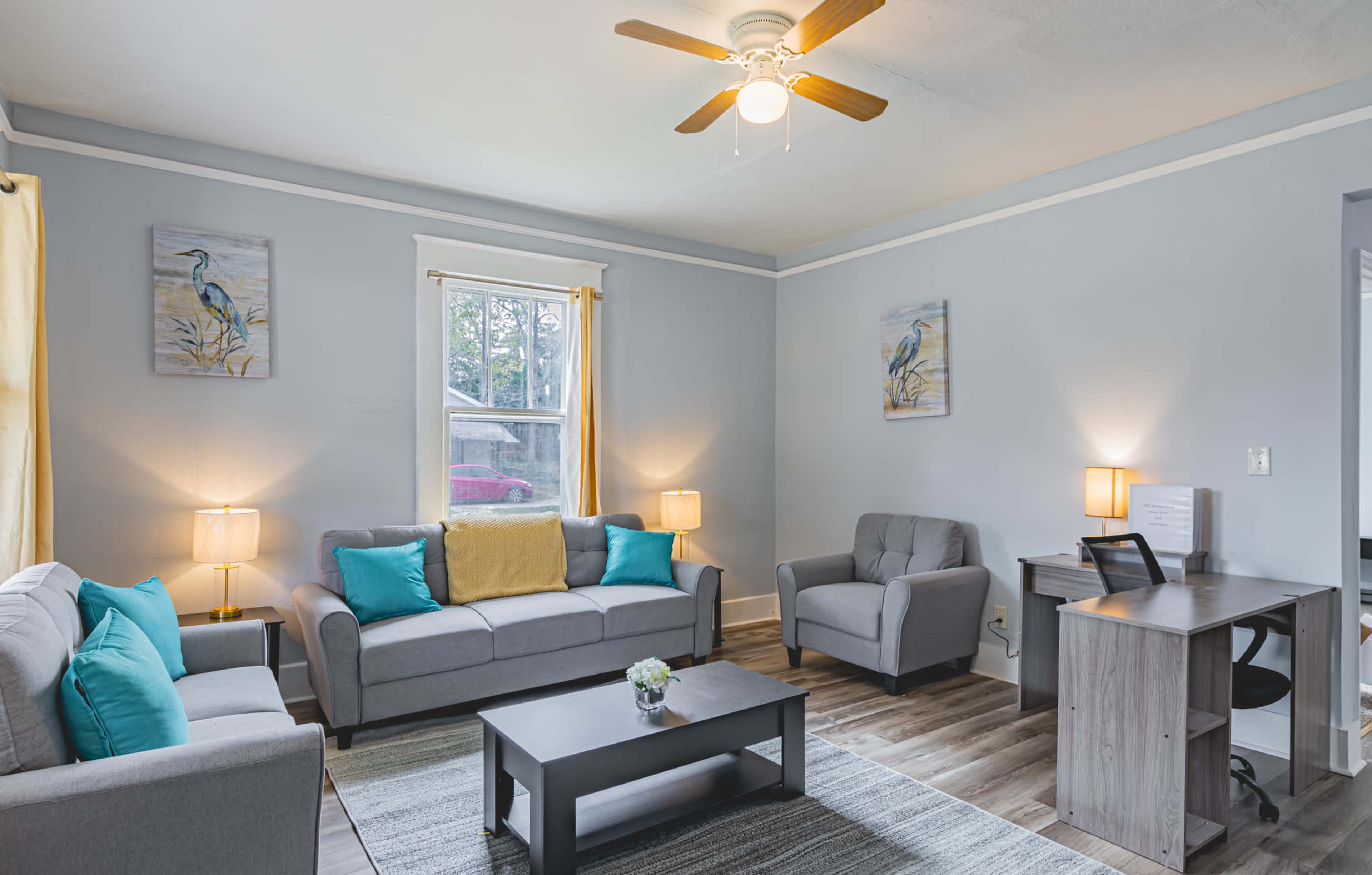 A modern living room features a gray couch, armchairs, a coffee table, and a desk, with yellow curtains and paintings of birds on the walls.
