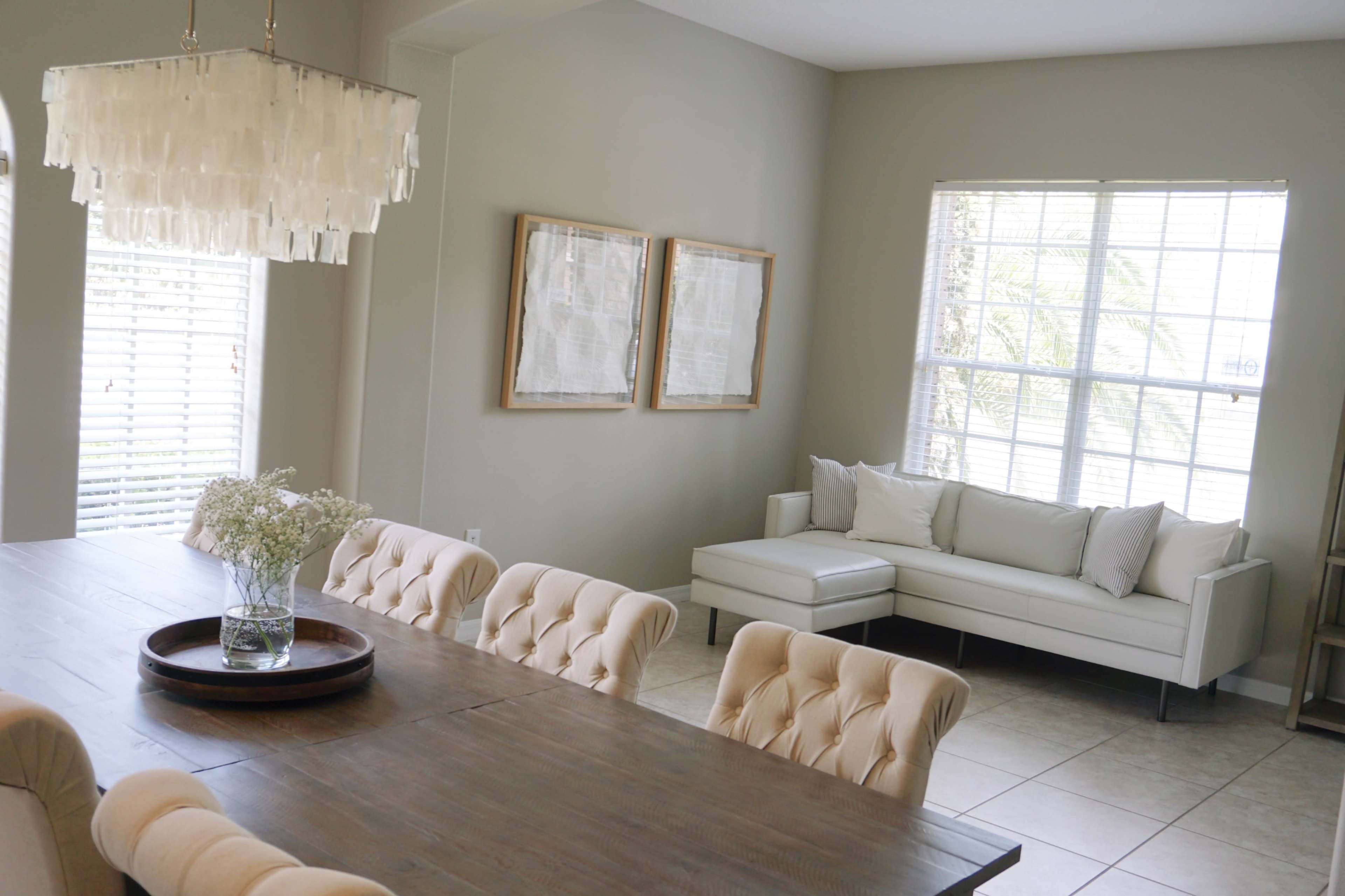A dining area features a long wooden table with tufted chairs, a chandelier above, and a light-filled corner with a white sofa and framed artwork on the walls.