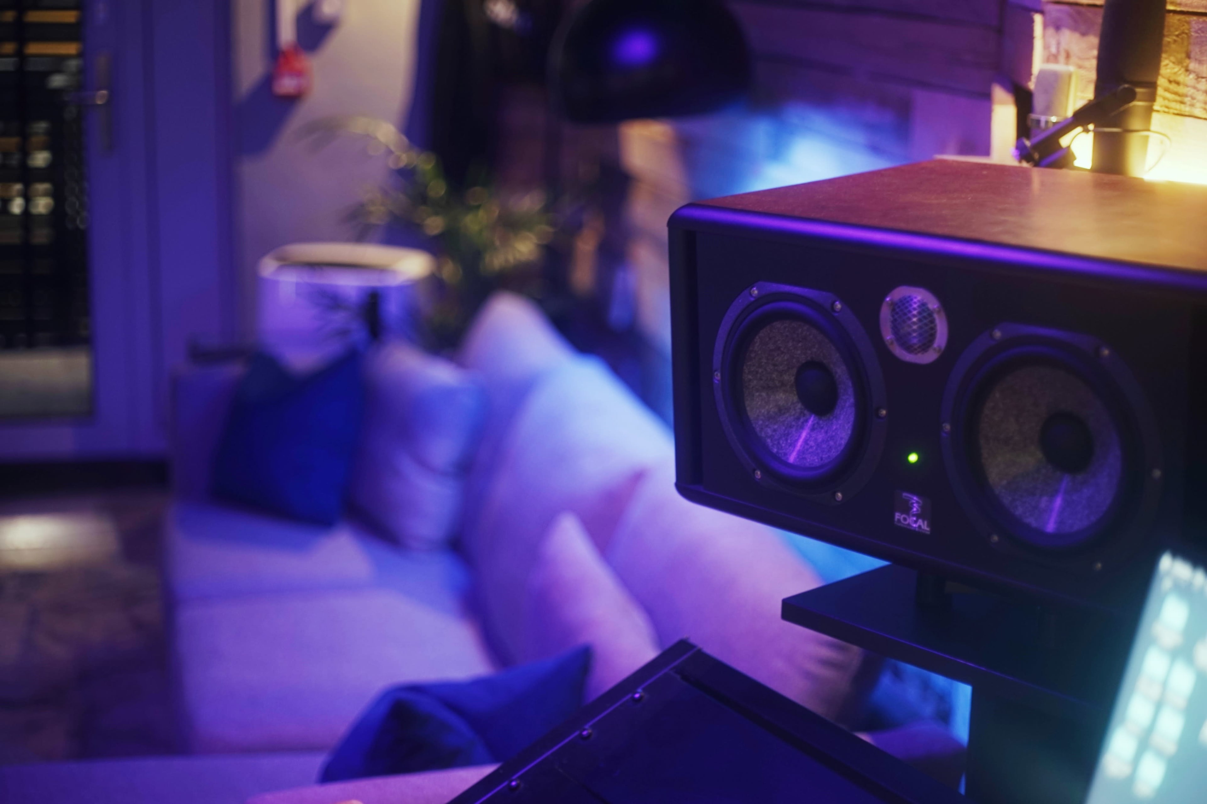 A pair of speakers is positioned on a shelf next to a couch in a dimly lit room with colorful lighting.