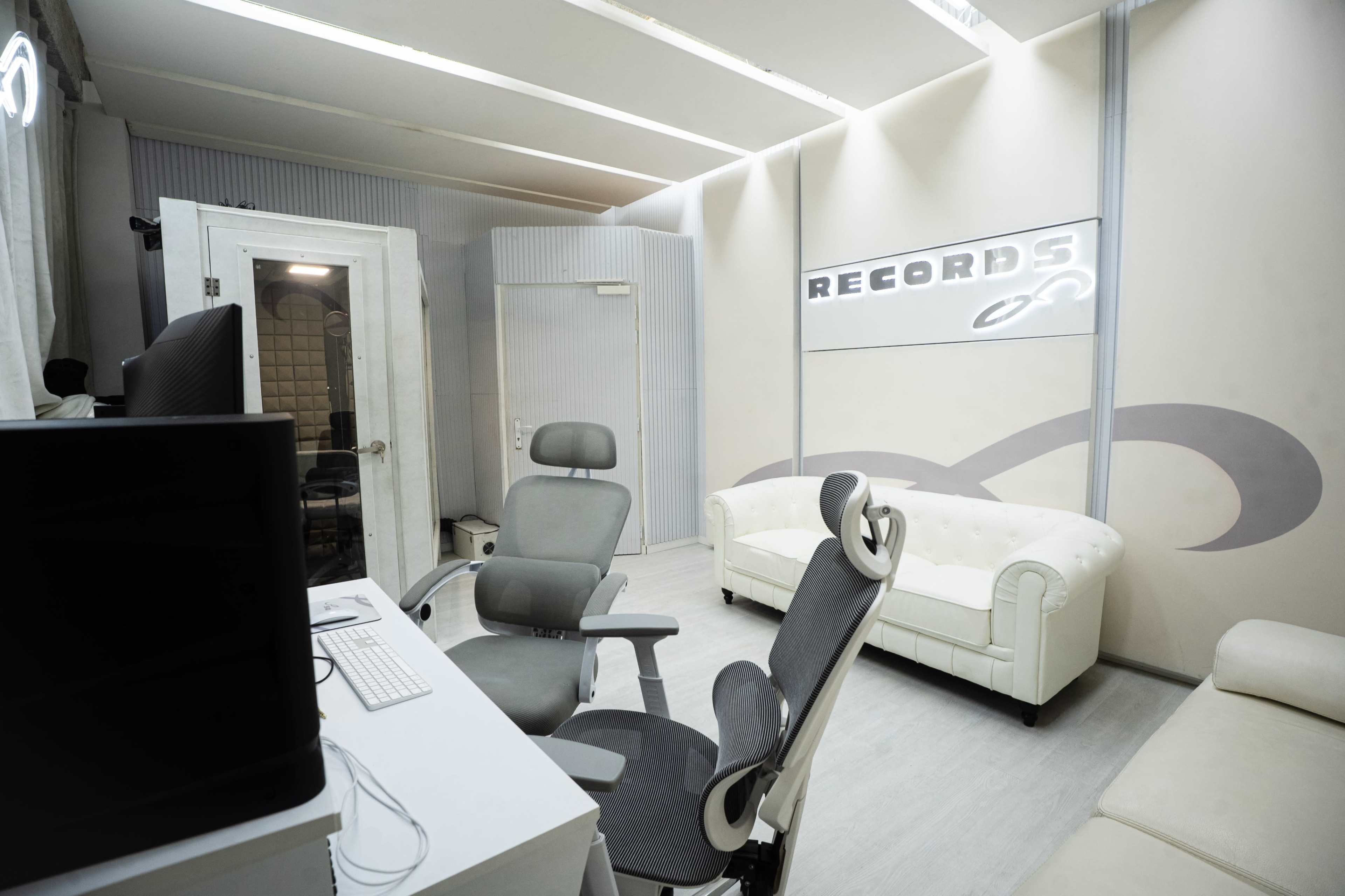 The image shows a modern recording studio with a white sofa, office chairs, and a computer setup against light-colored walls featuring a "RECORDS" sign.