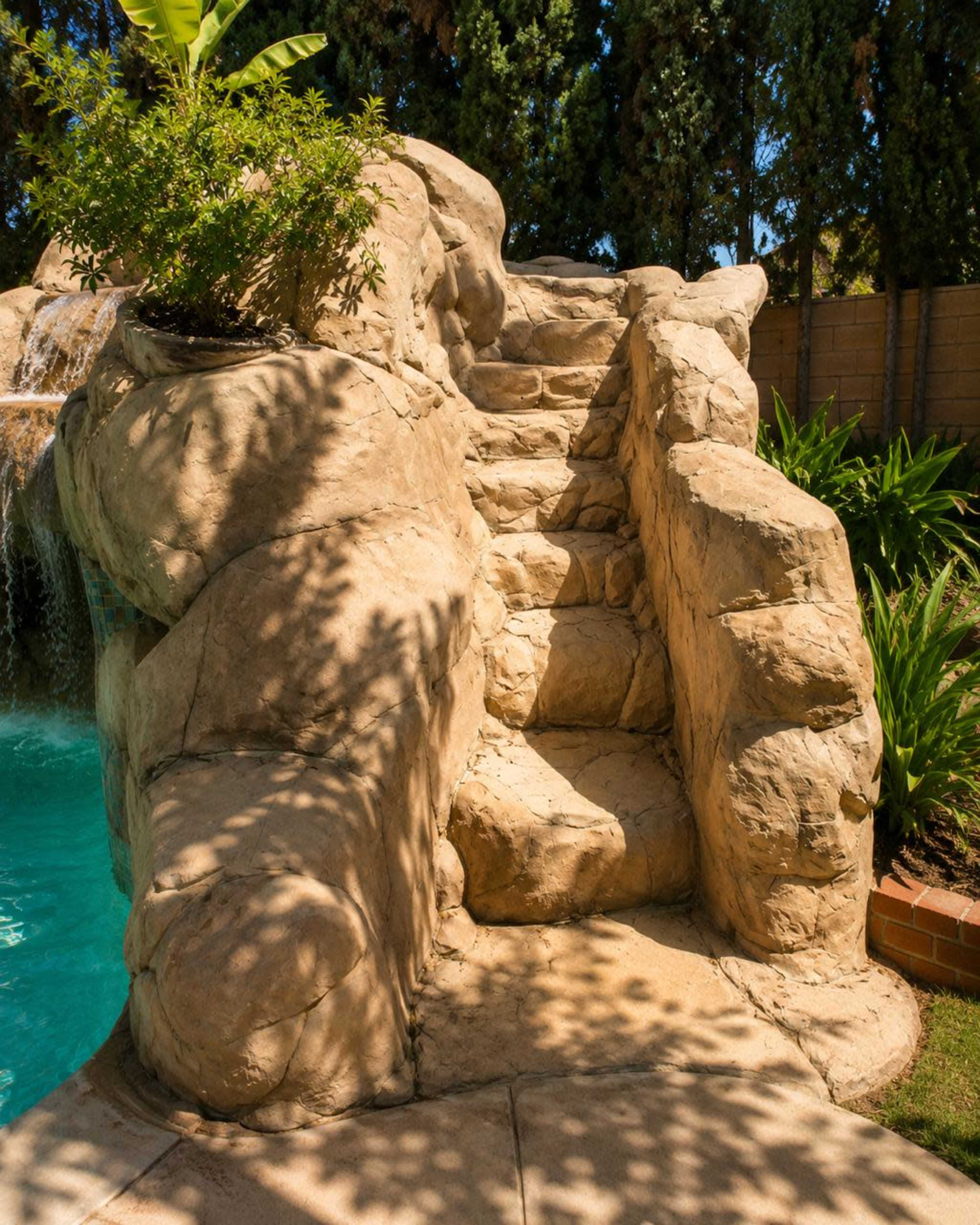 The image shows a rock-like water slide with steps leading down to a swimming pool, surrounded by grass and plants.