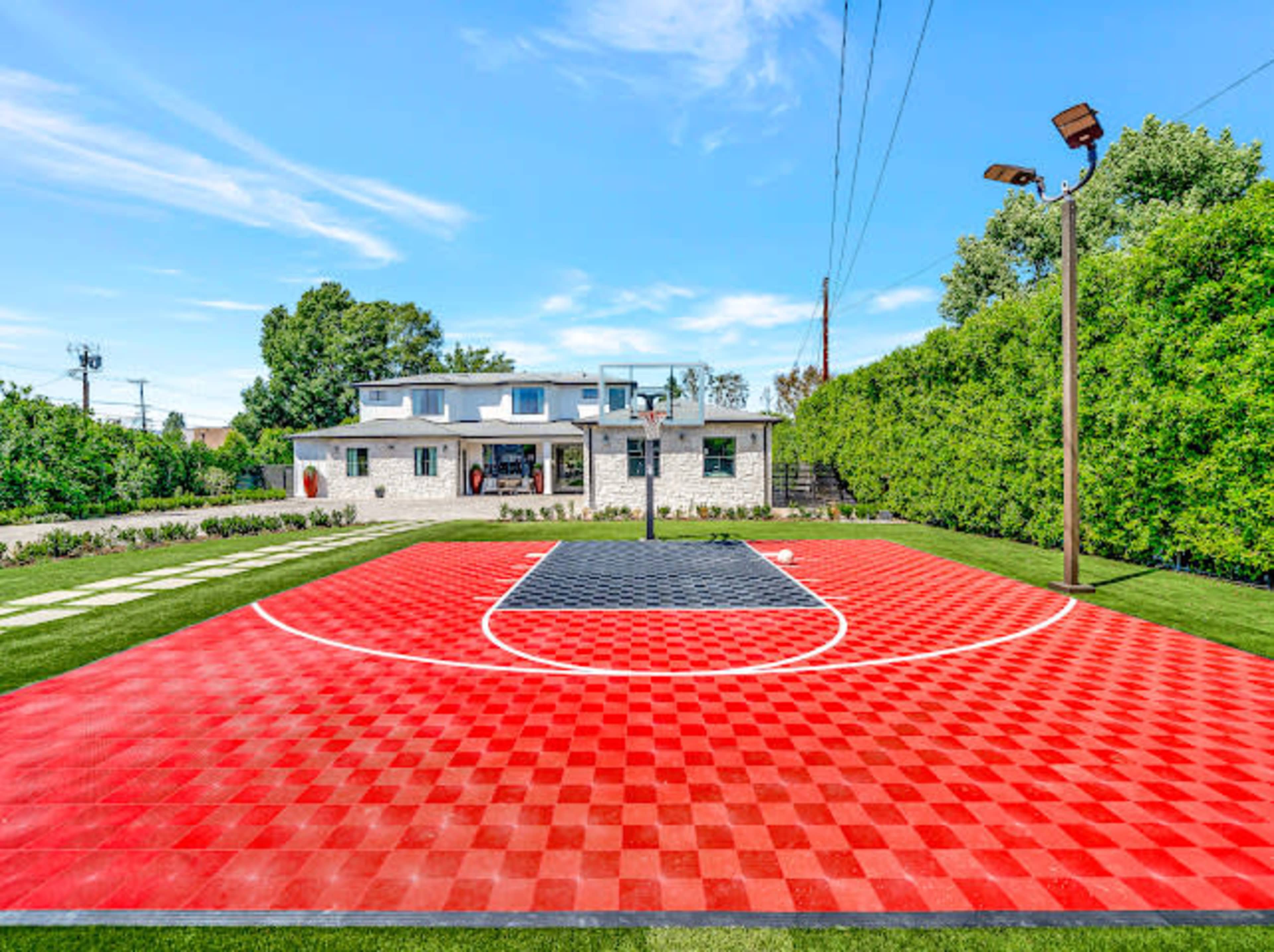 The image shows a vibrant red and black outdoor basketball court with a house and greenery in the background.