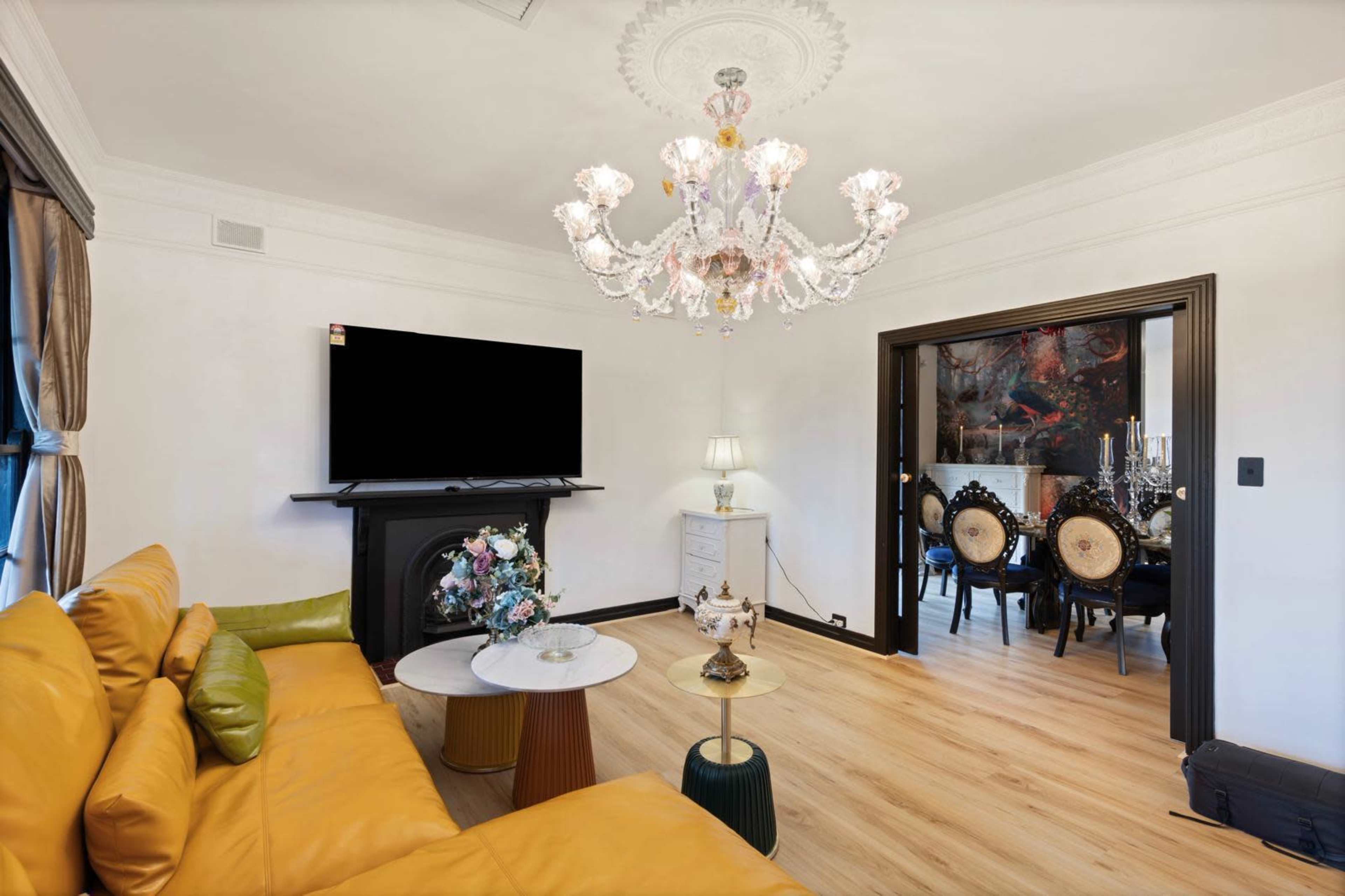 The image shows a living room with a yellow sofa, a chandelier, a television mounted above a fireplace, and a dining area with ornate chairs in the background.