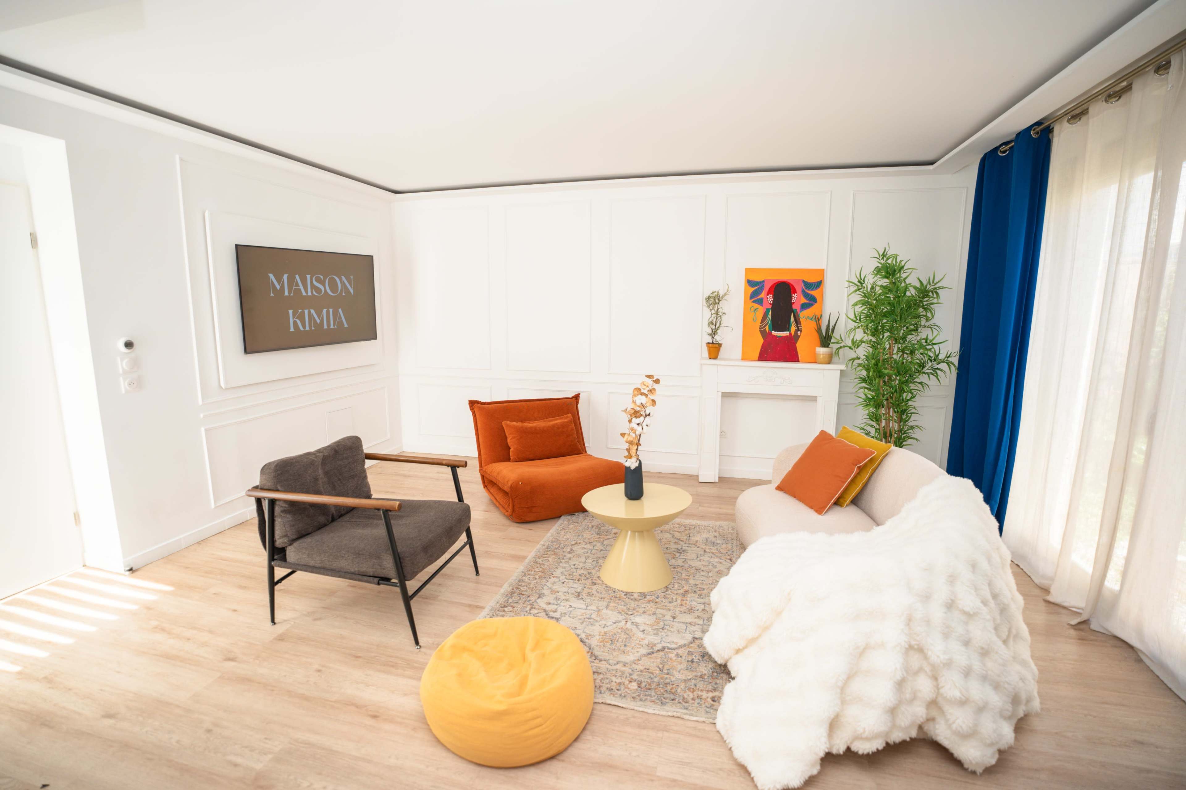 The image shows a brightly lit living room with a mix of modern furniture, including a gray chair, an orange armchair, a white sofa, and a round table, surrounded by decorative plants and a colorful artwork on the wall.