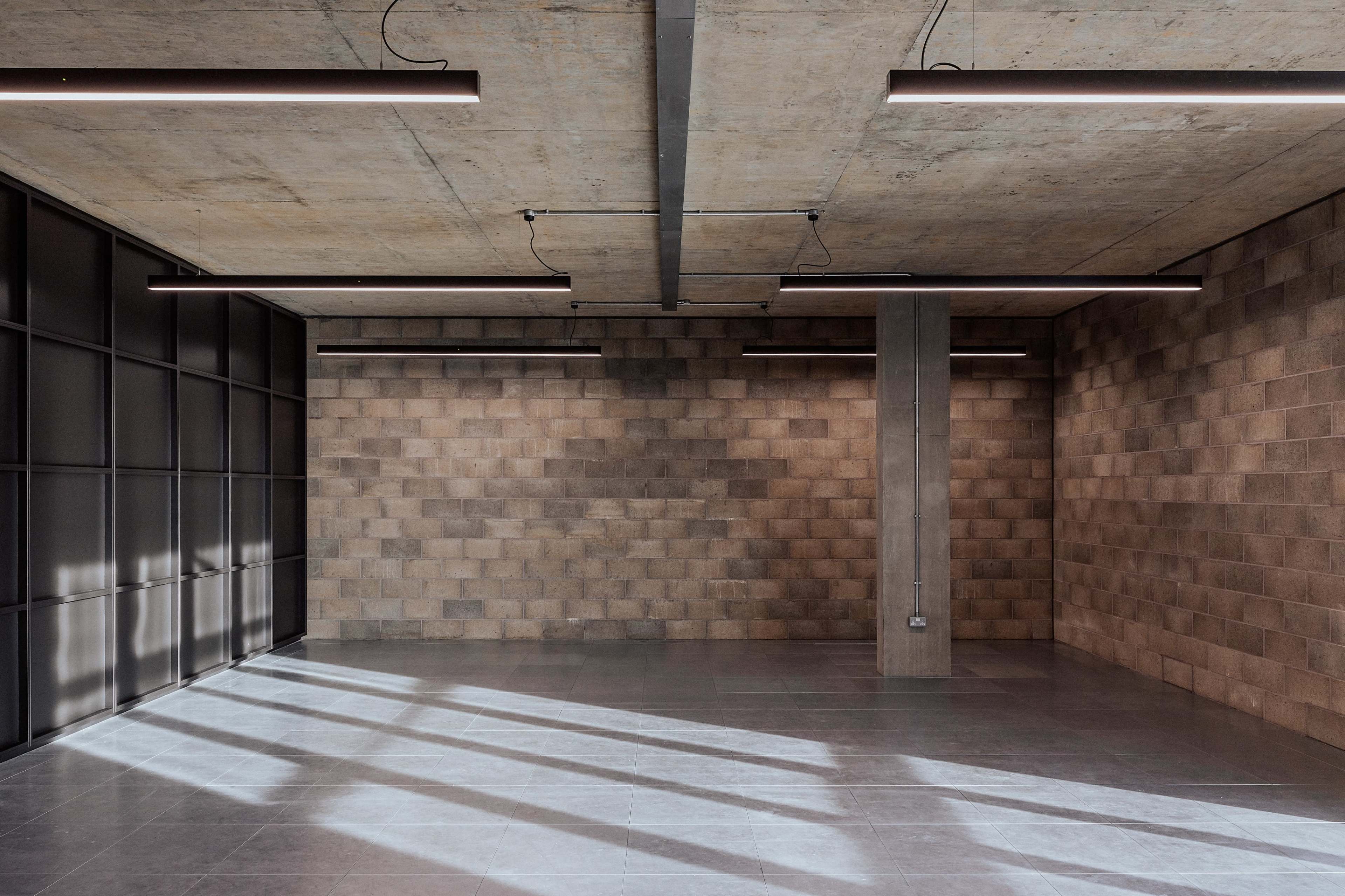 The image shows a spacious interior room with concrete walls and a polished floor, featuring modern lighting fixtures overhead and large windows casting shadows on the floor.