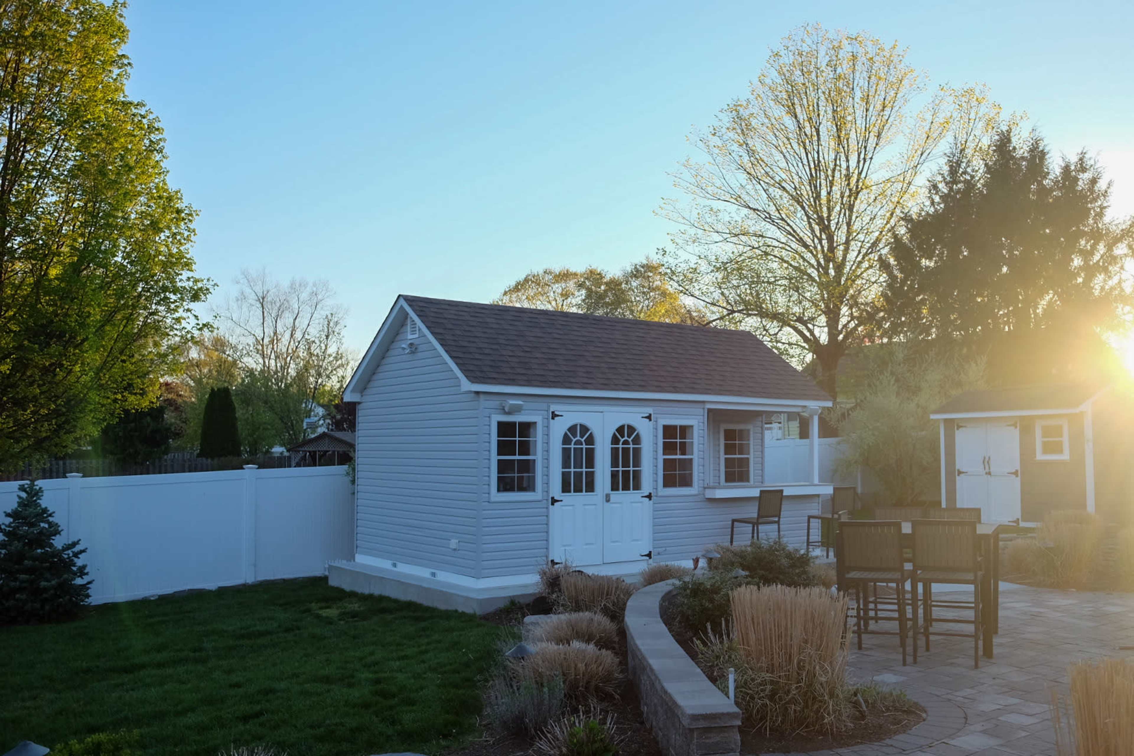 A small white shed with a shingled roof is located beside a stone patio and surrounded by greenery in a backyard at sunset.