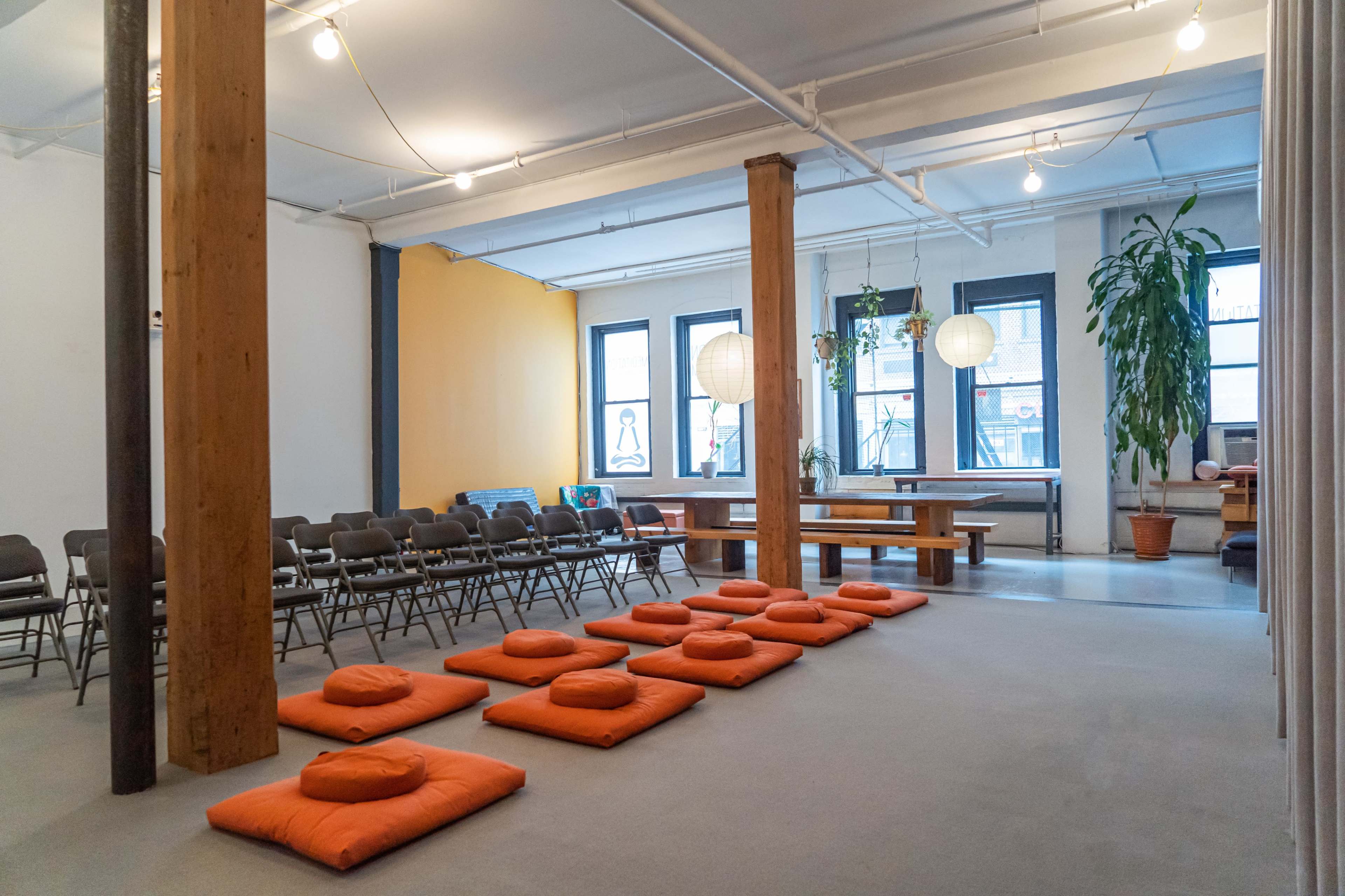 A spacious room with orange floor cushions arranged on a carpet, folding chairs set in rows, and large windows allowing natural light to enter.