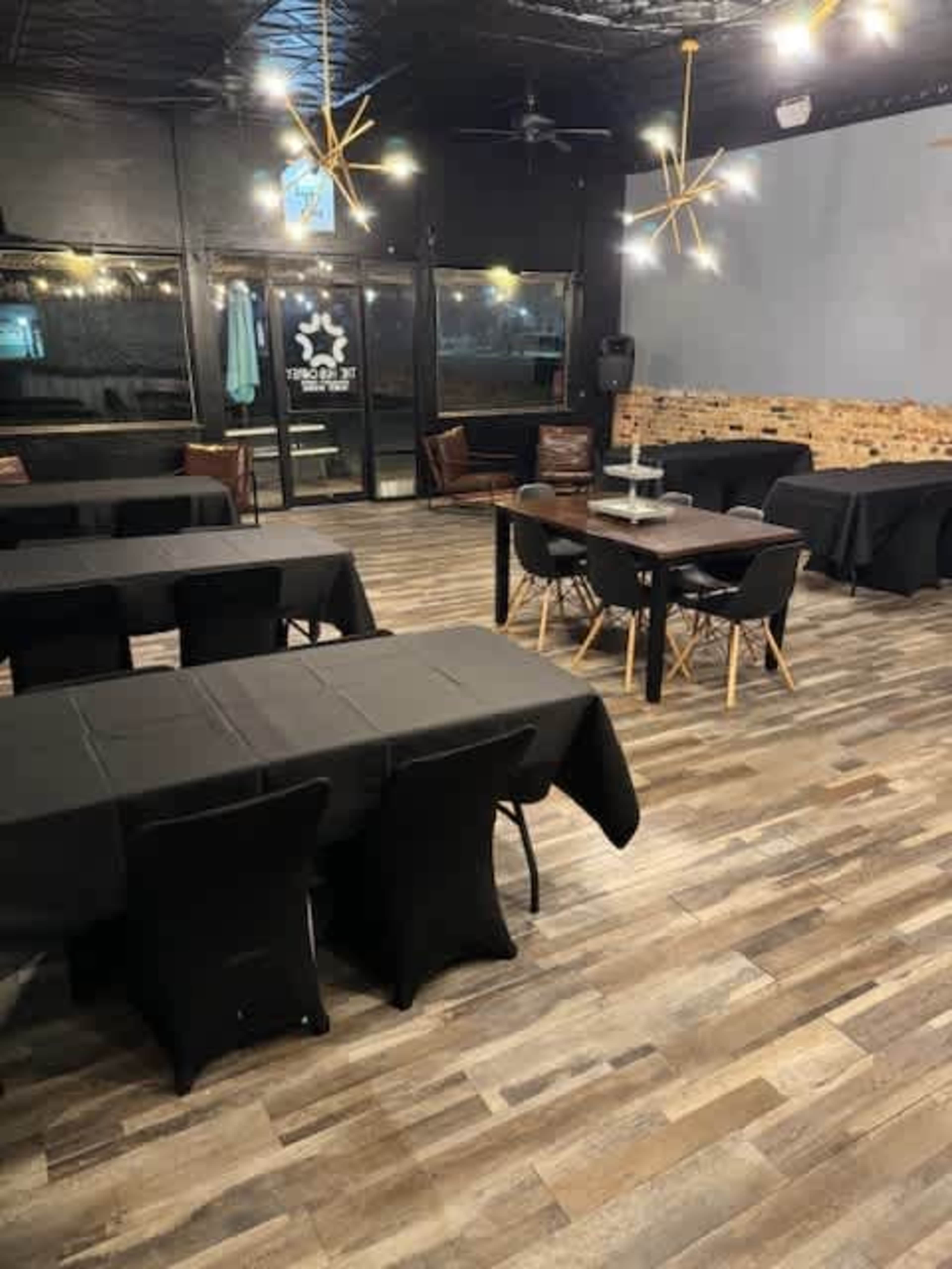 The image shows an empty dining area with black tablecloths covering tables, wooden flooring, and modern lighting fixtures overhead.