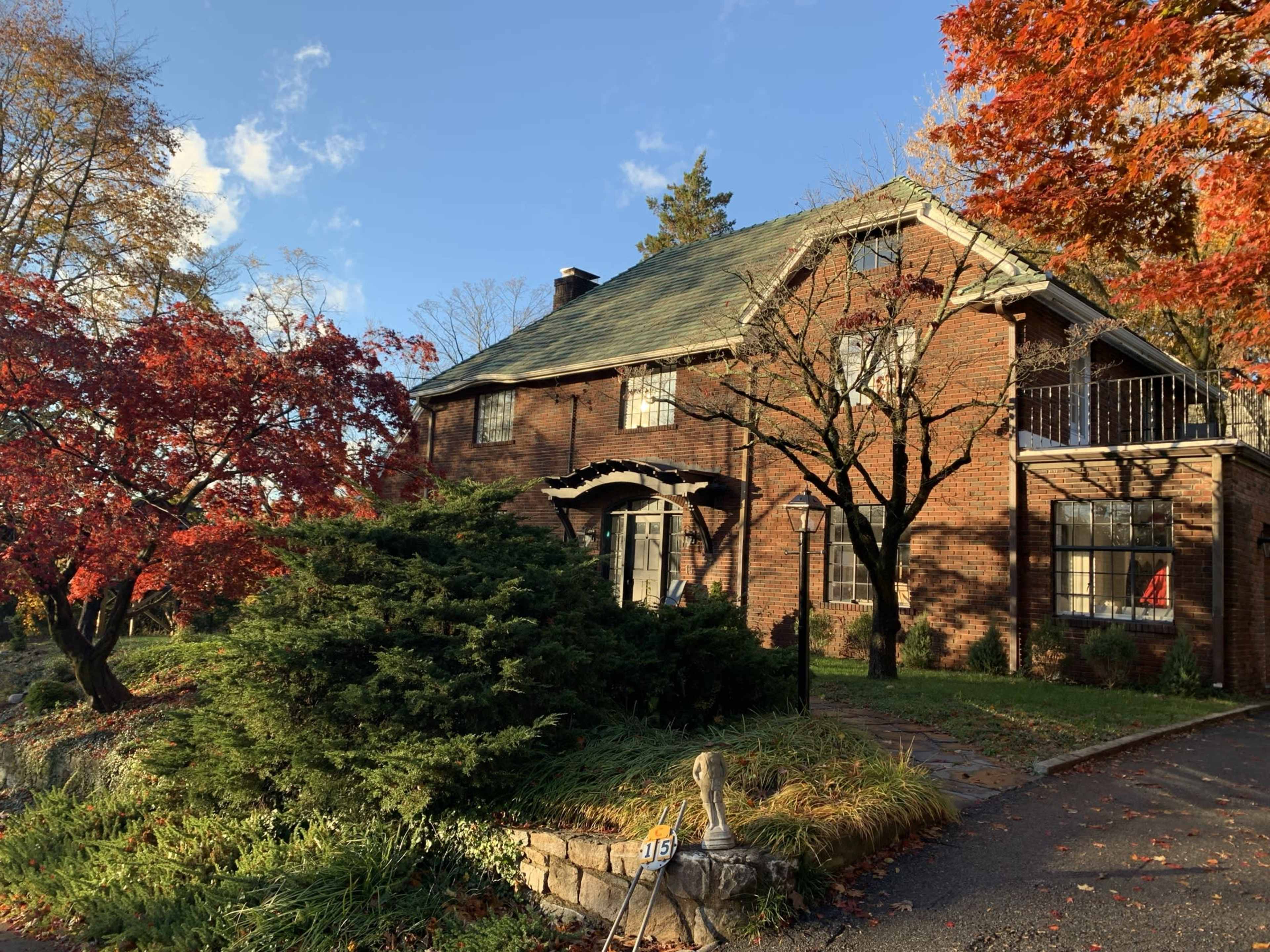 A brick house with a sloped roof is surrounded by colorful autumn foliage and greenery.