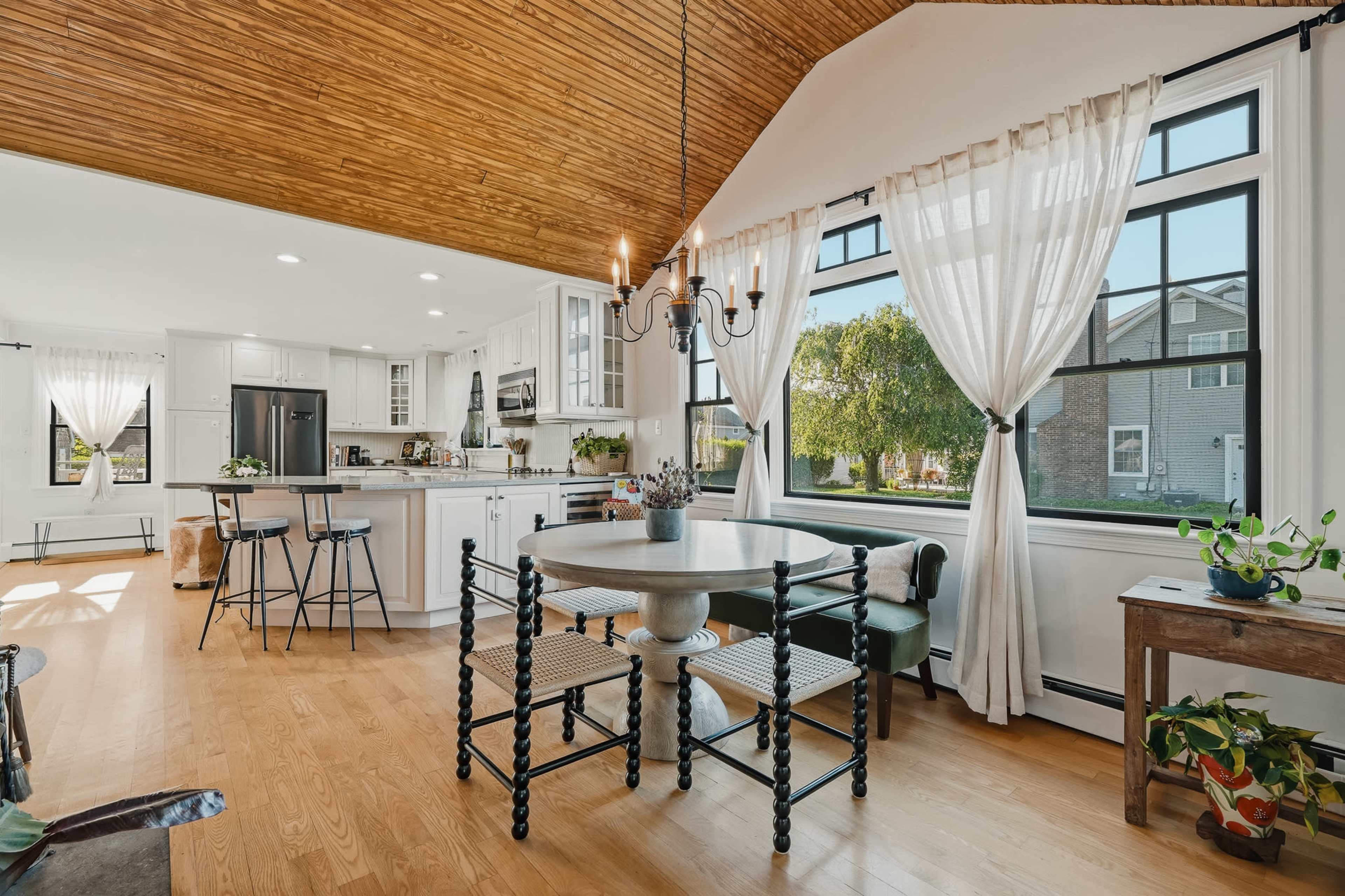 The image shows a bright kitchen and dining area with white cabinetry, a round table, and large windows adorned with sheer curtains.