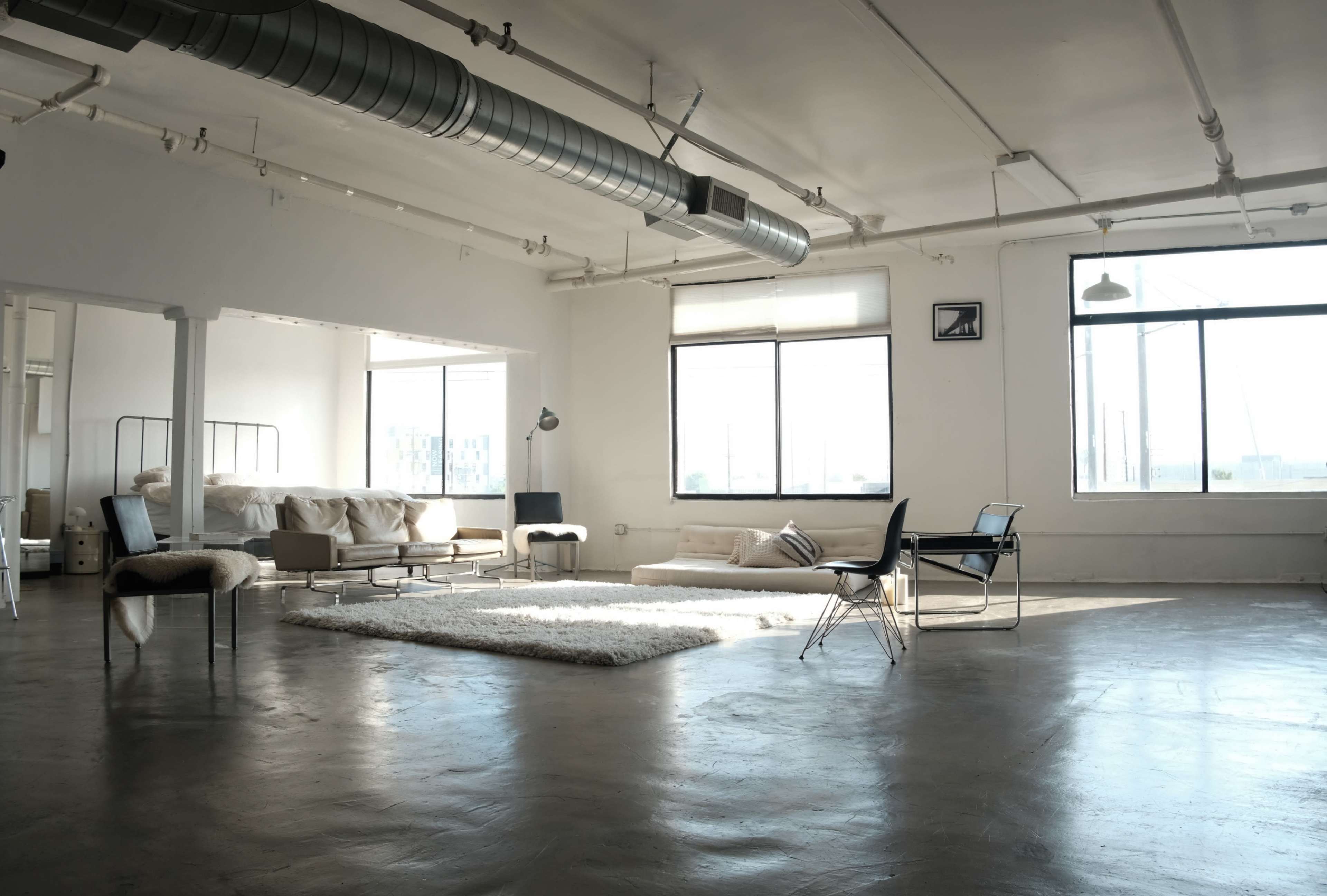 The image shows a spacious, minimalist studio apartment with large windows, industrial features, and simple furniture including a bed, couch, and a rug.