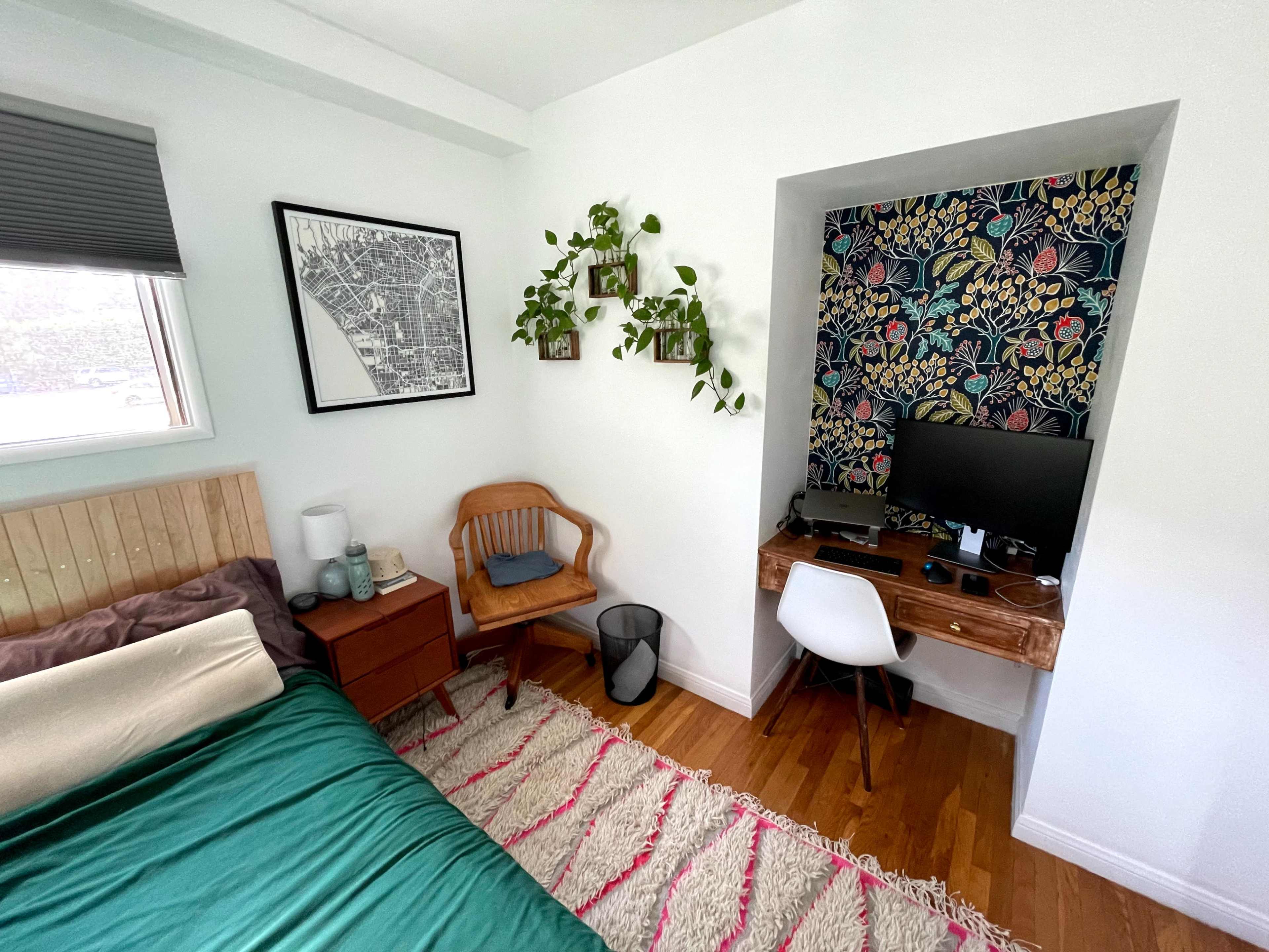 A modern bedroom features a wooden bed, a desk with a monitor, and a vibrant floral accent wall.
