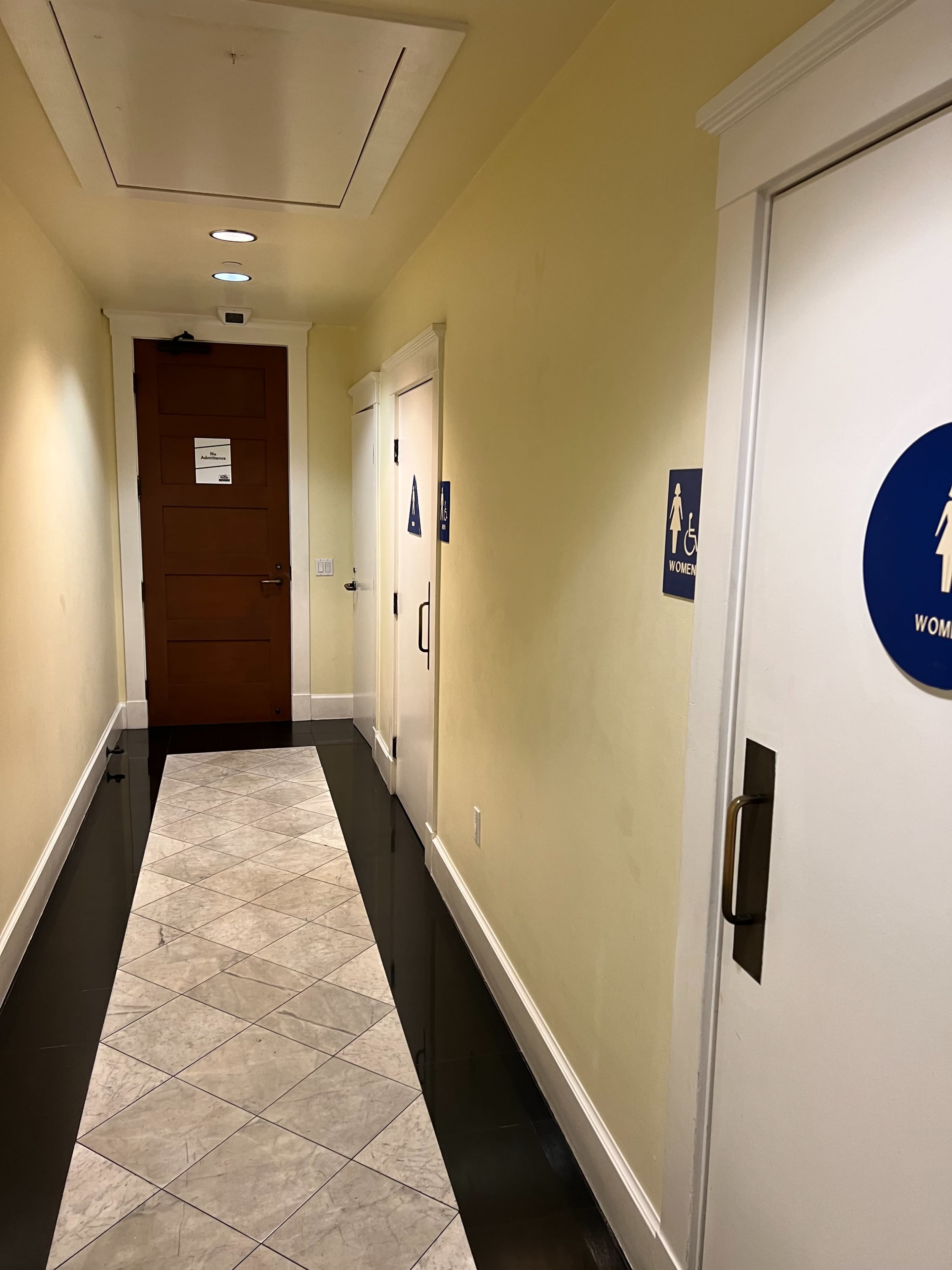 A narrow hallway with a tiled floor leading to two restroom doors labeled "WOMEN" and a sign on the door at the end.