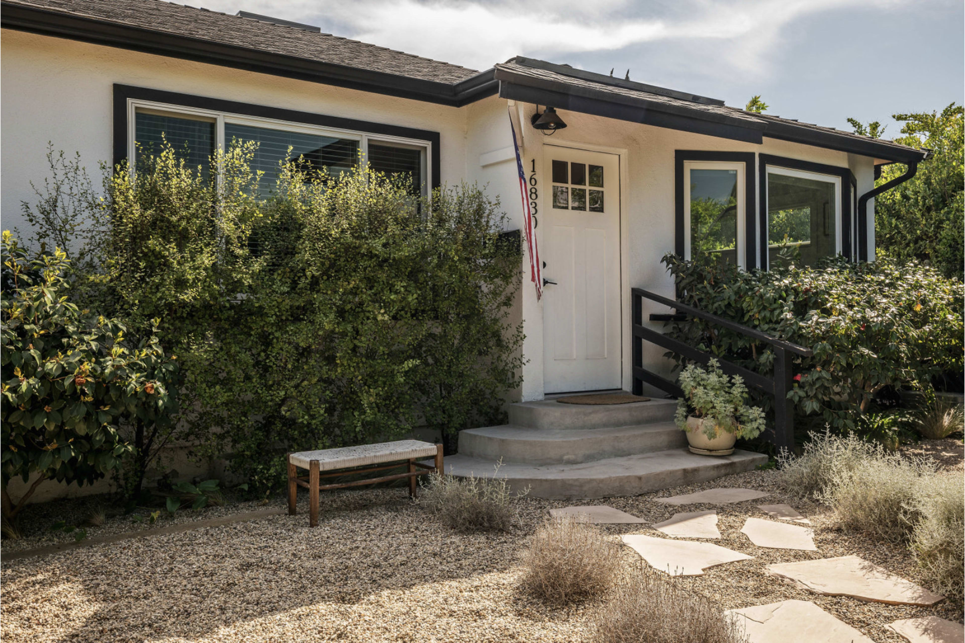 The image shows a house with a white exterior, a front door with a small porch, and a landscaped yard featuring various plants and gravel pathways.