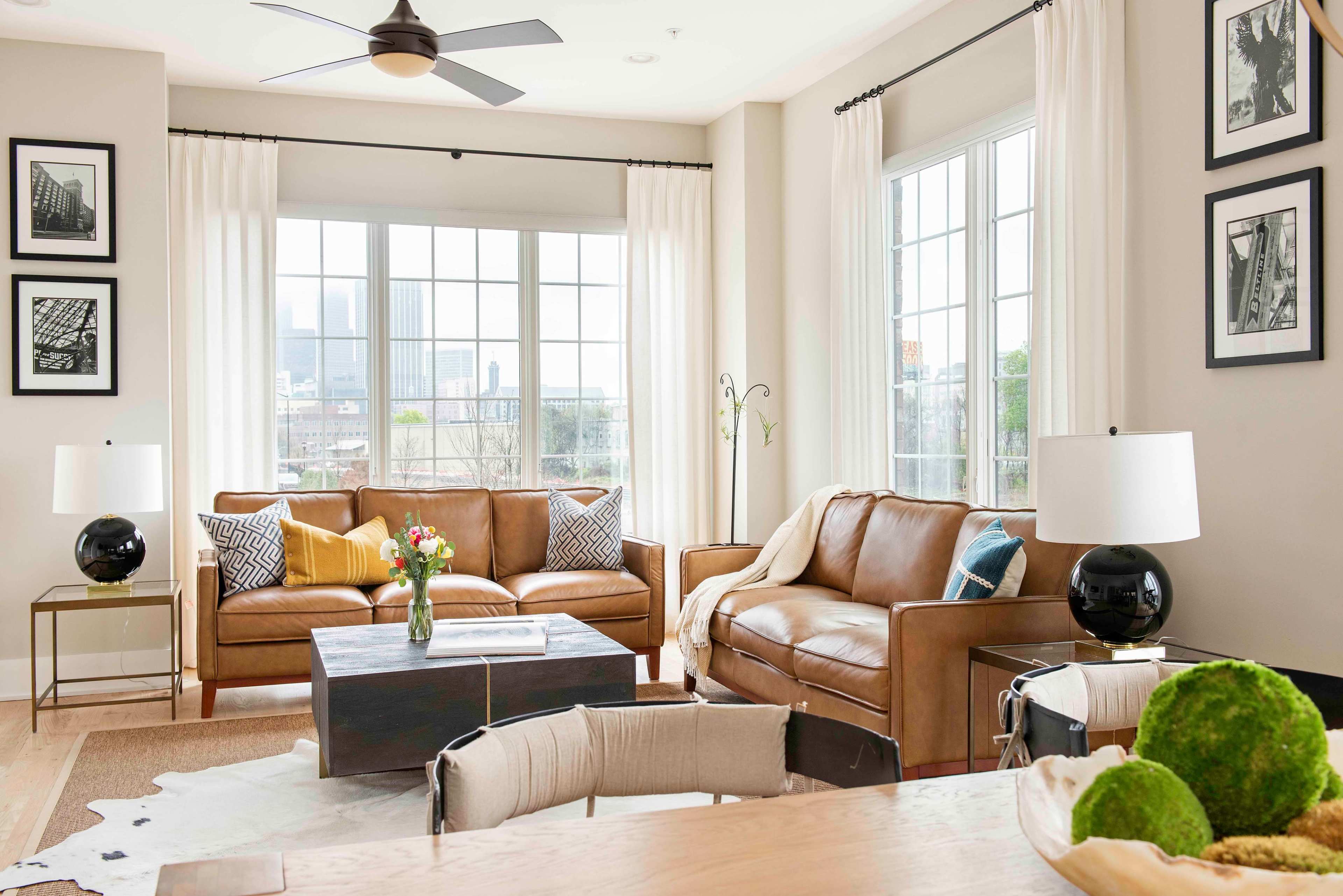The image shows a bright living room with large windows, a leather sofa set, a coffee table, and decor featuring black and white photographs.