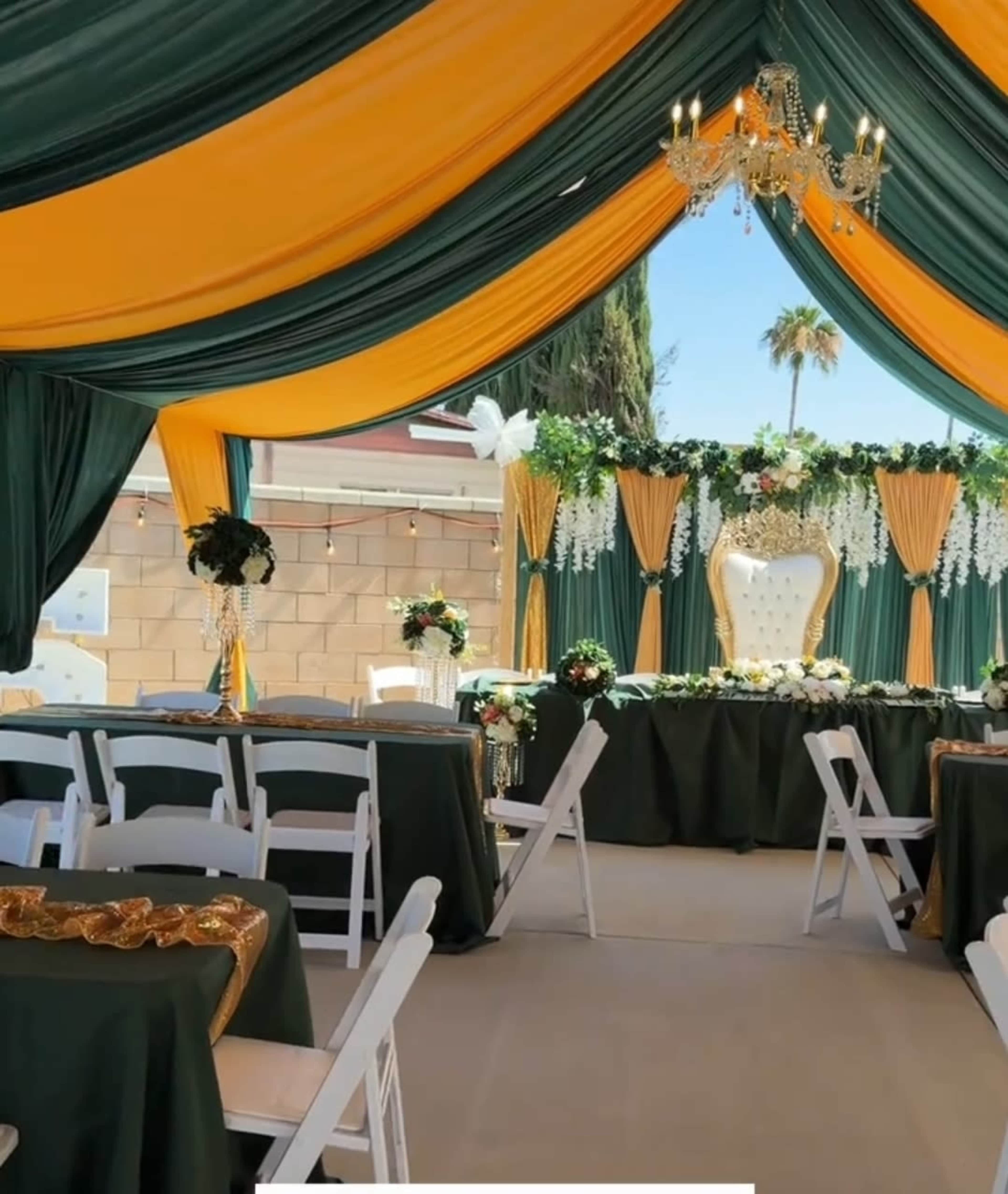 The image depicts a beautifully decorated event space with a tent featuring green and gold draping, a floral backdrop, and elegantly arranged tables and chairs.