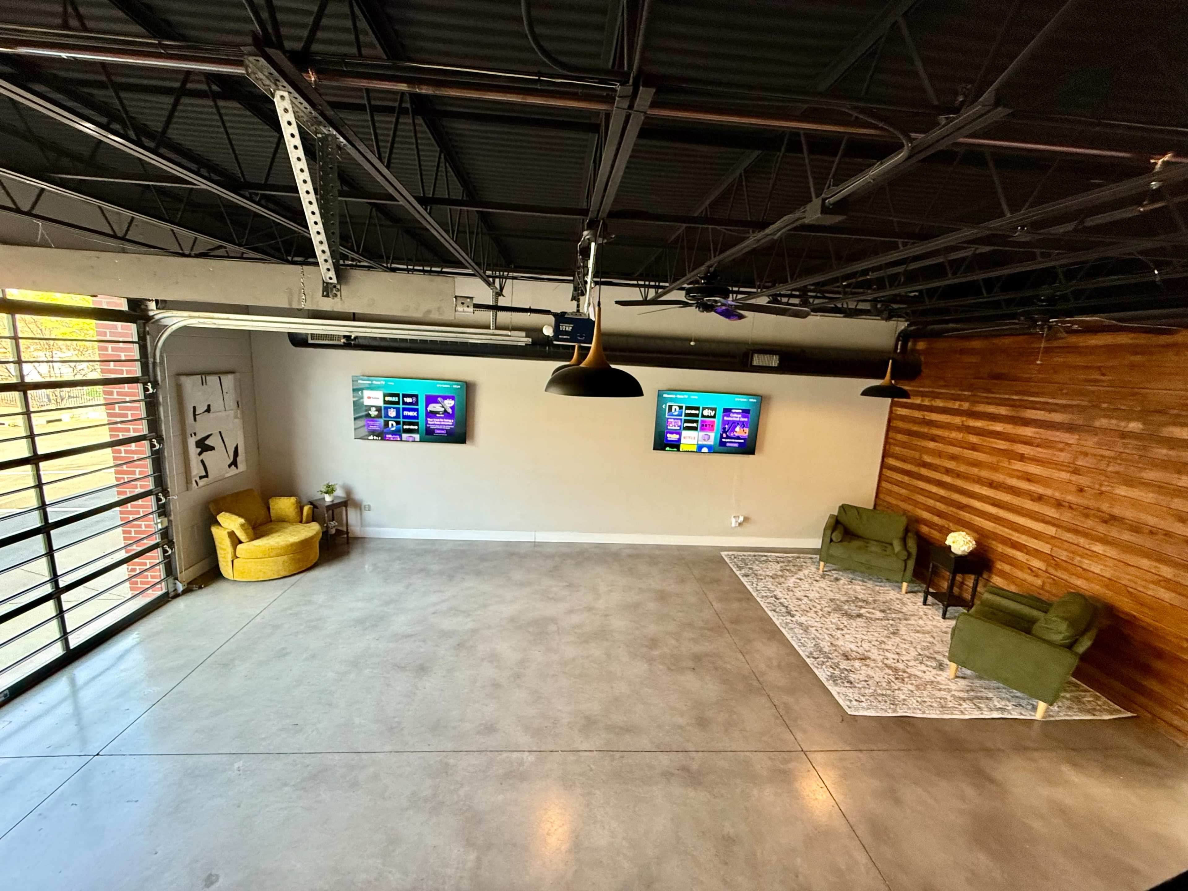The image shows a spacious room with a concrete floor, featuring two green chairs and two wall-mounted screens on one side, while the other side has a wooden wall and large windows.