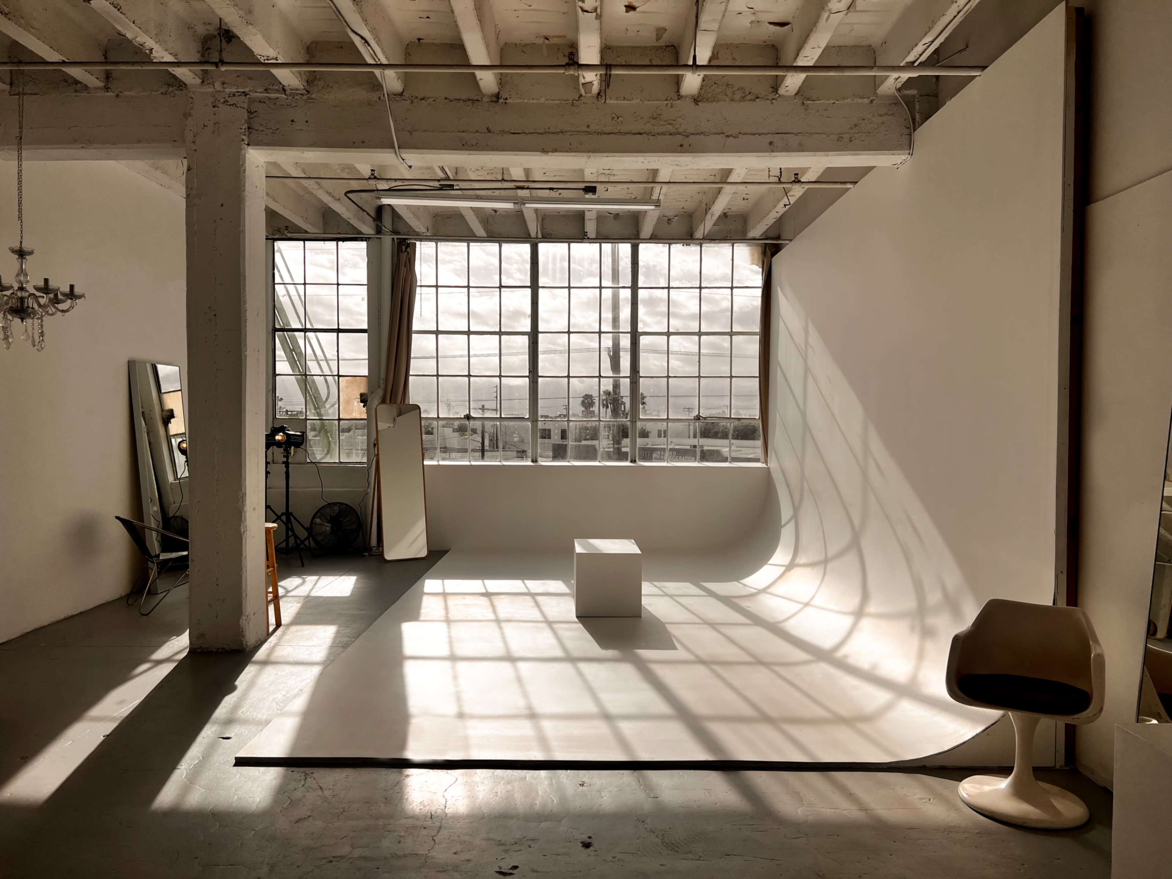 A photography studio features a large window allowing natural light to cast shadows on a seamless backdrop, with a white cube in the center and a chair nearby.