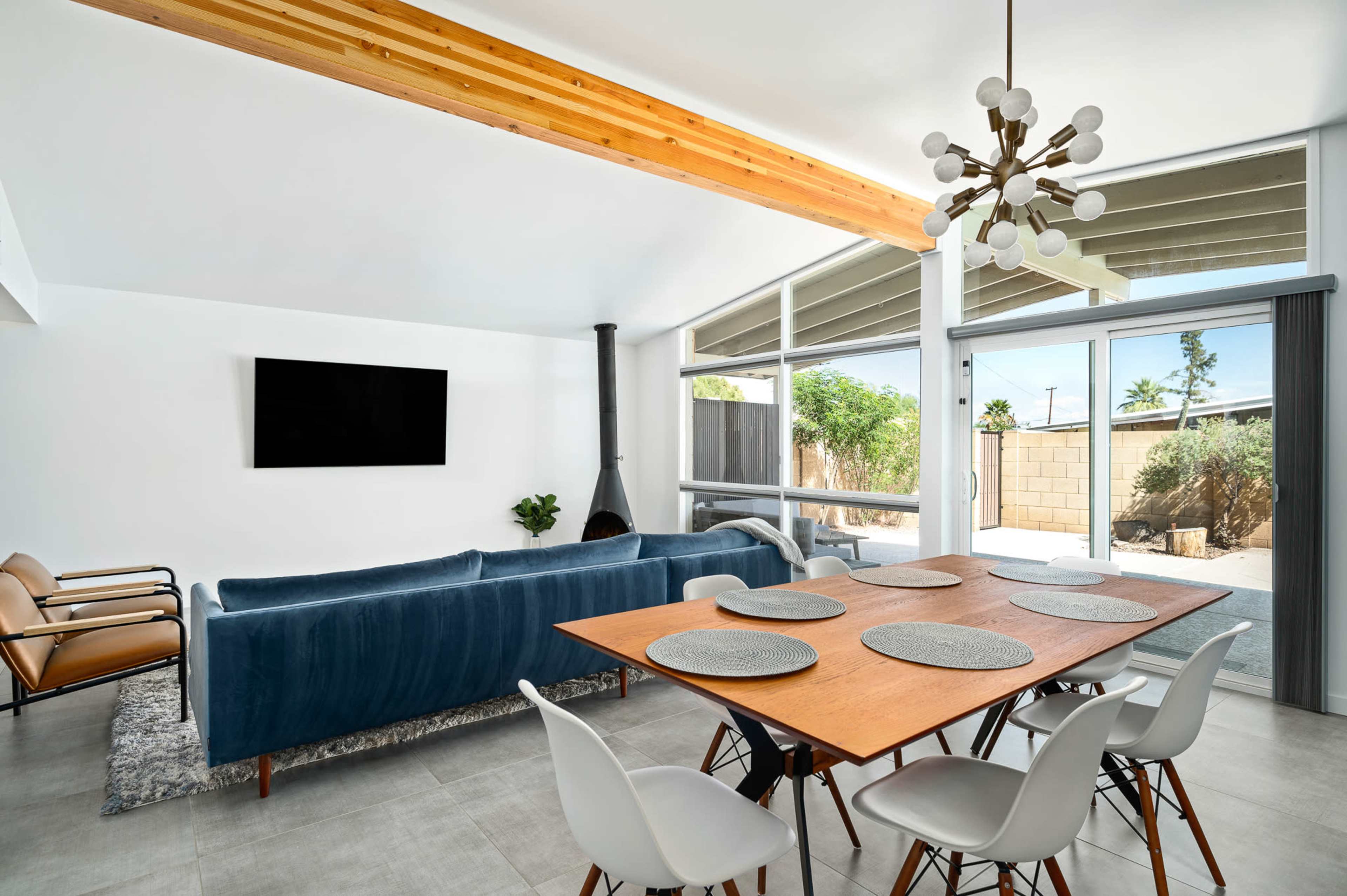 A modern living area features a blue sofa, a wooden dining table set for six, and large windows with a view of a patio garden.