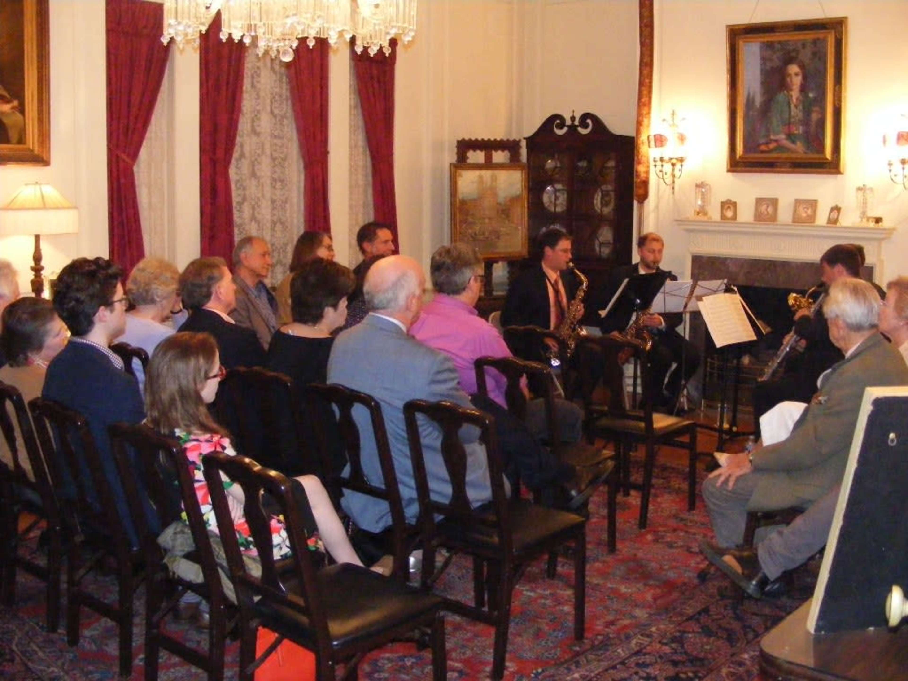 A group of people is seated in a room with elegant decor, watching a small musical performance by musicians positioned near a fireplace.
