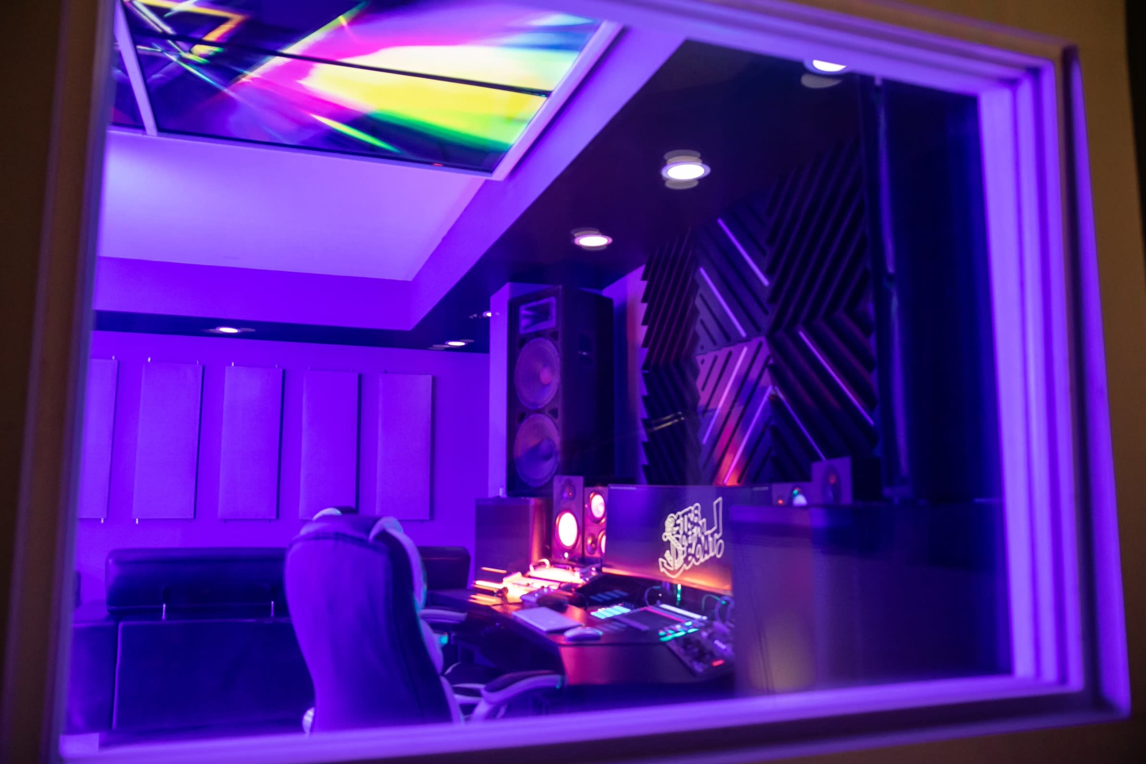 A music production studio featuring purple lighting, a desk with electronic equipment, and acoustic panels on the walls.