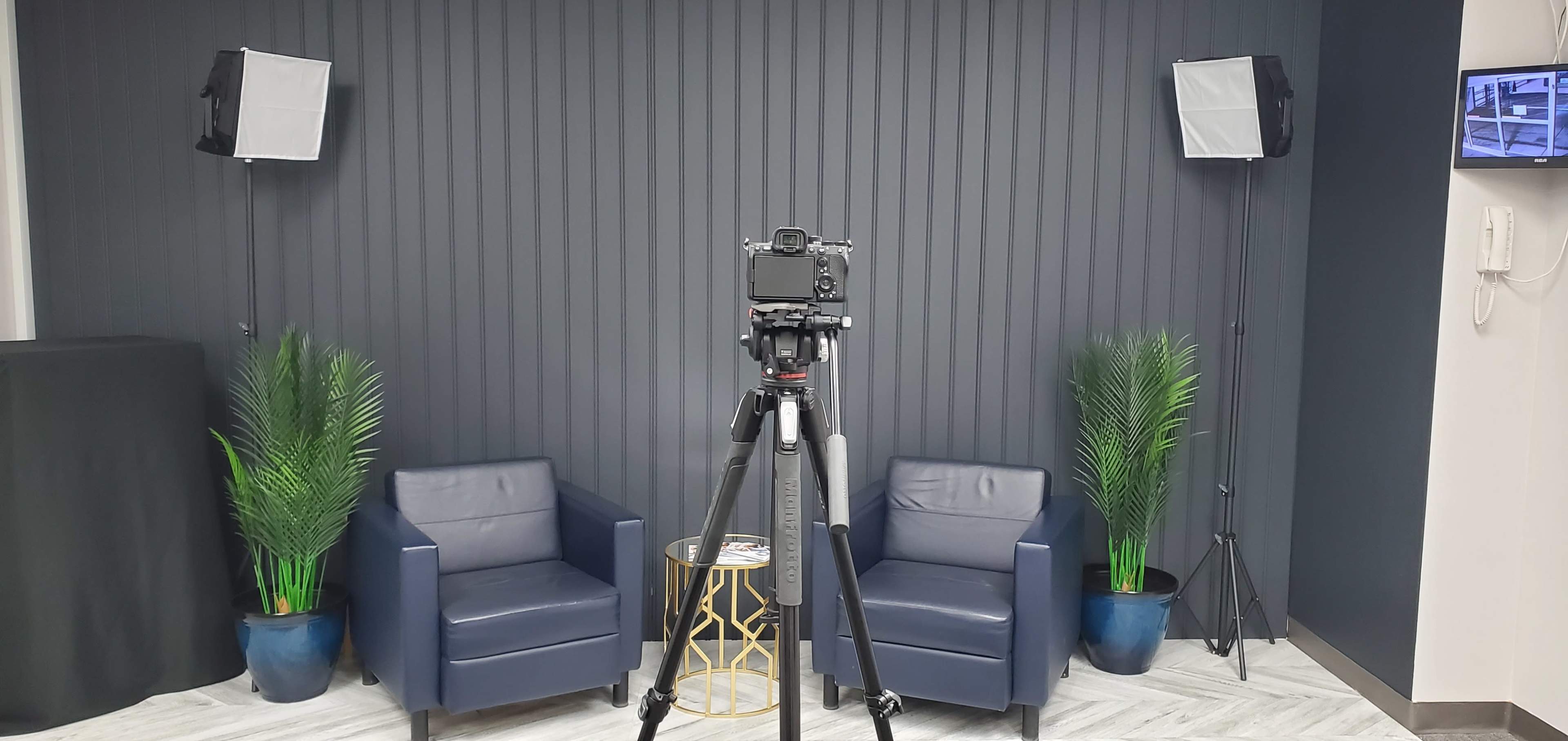 A setup with two blue chairs on either side of a small round table, a camera on a tripod facing the chairs, and lighting equipment on either side against a dark striped wall.