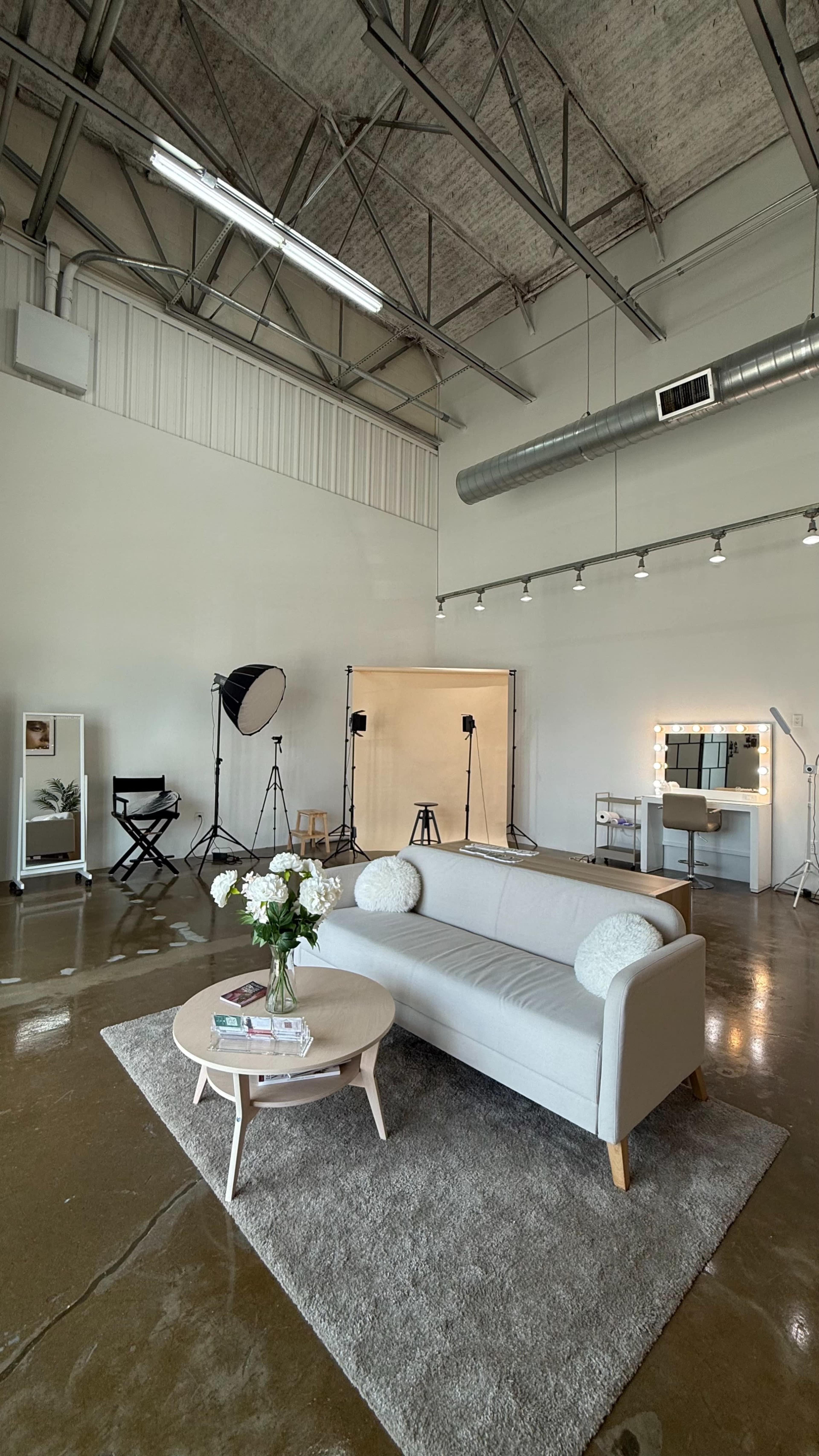 The image shows a spacious studio with a gray couch, a round coffee table with flowers, and various lighting and photography equipment set up against a plain backdrop.