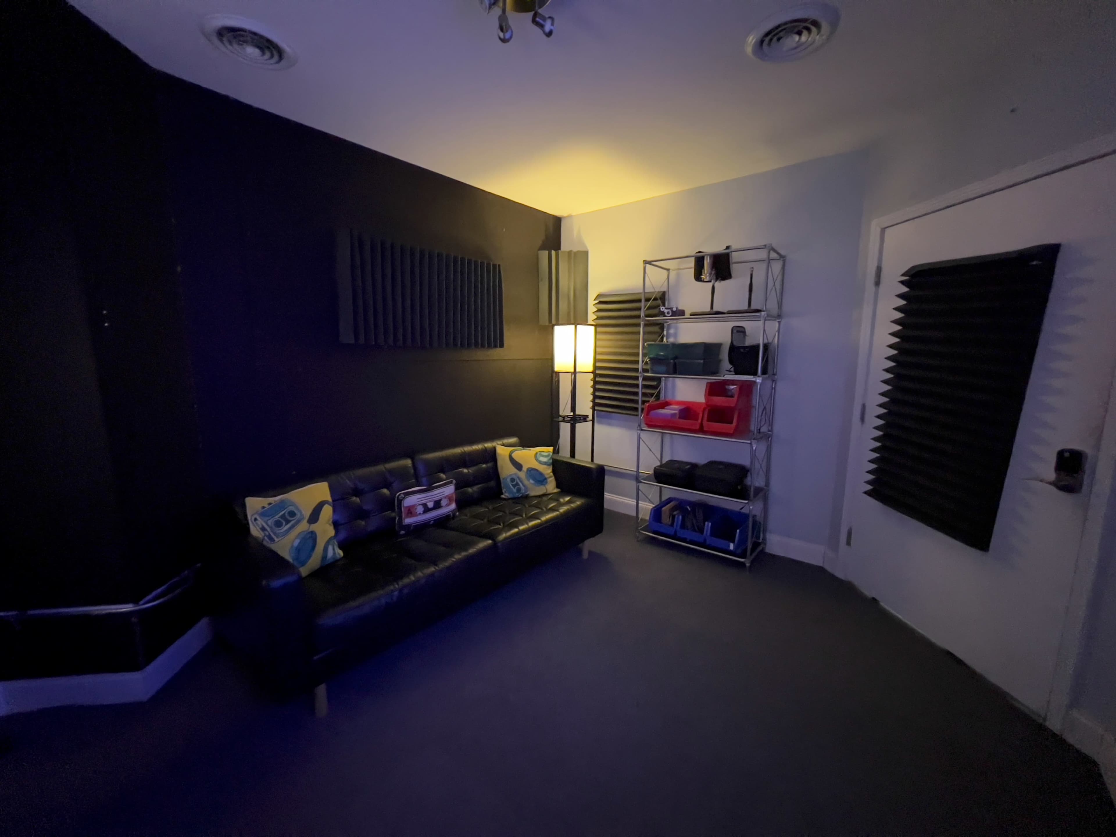 The image shows a dark room with a black wall, a couch with patterned cushions, a lamp, and a wire shelf containing various items.