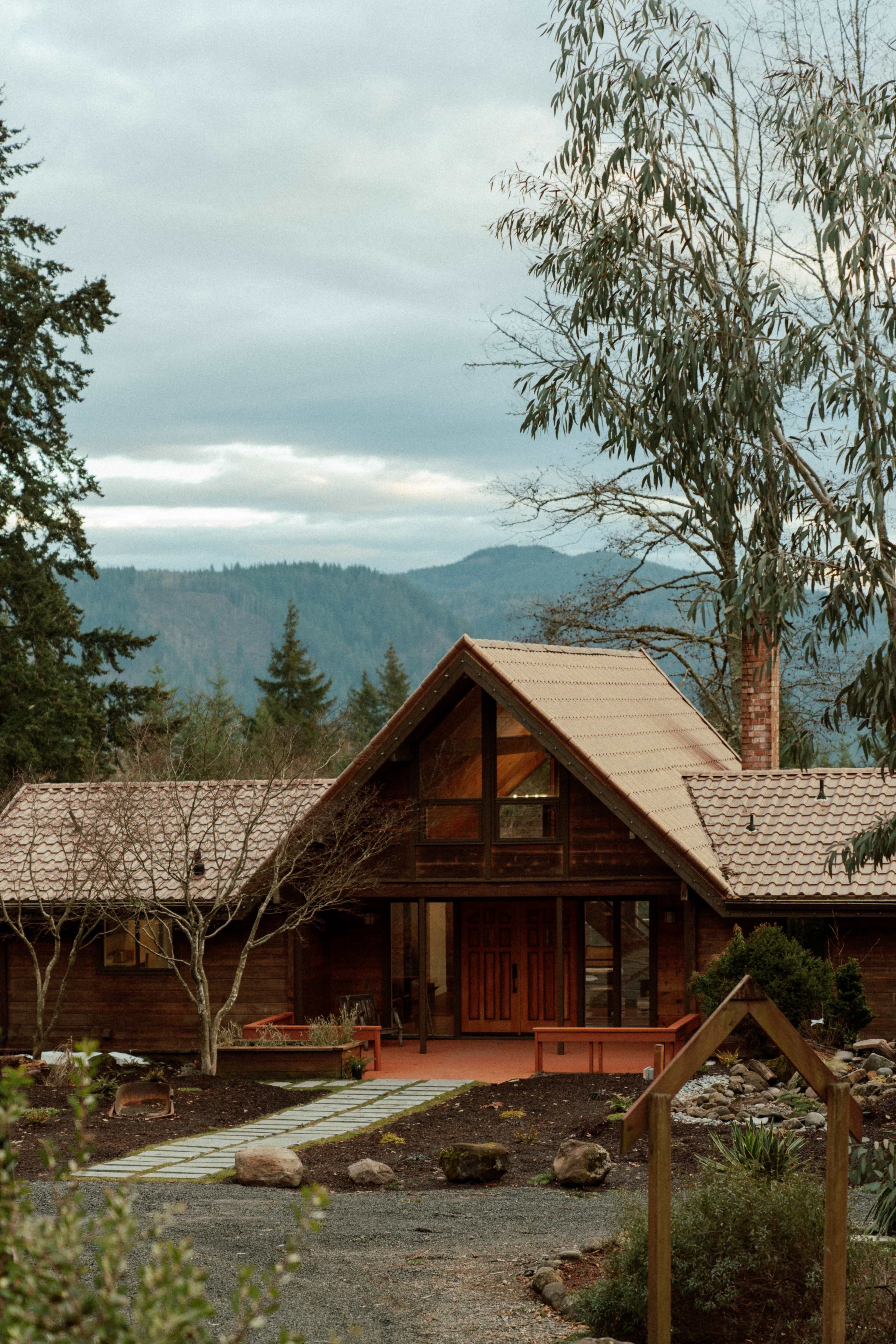 A wooden cabin with a sloped roof and large front windows is situated in a mountainous landscape surrounded by trees.