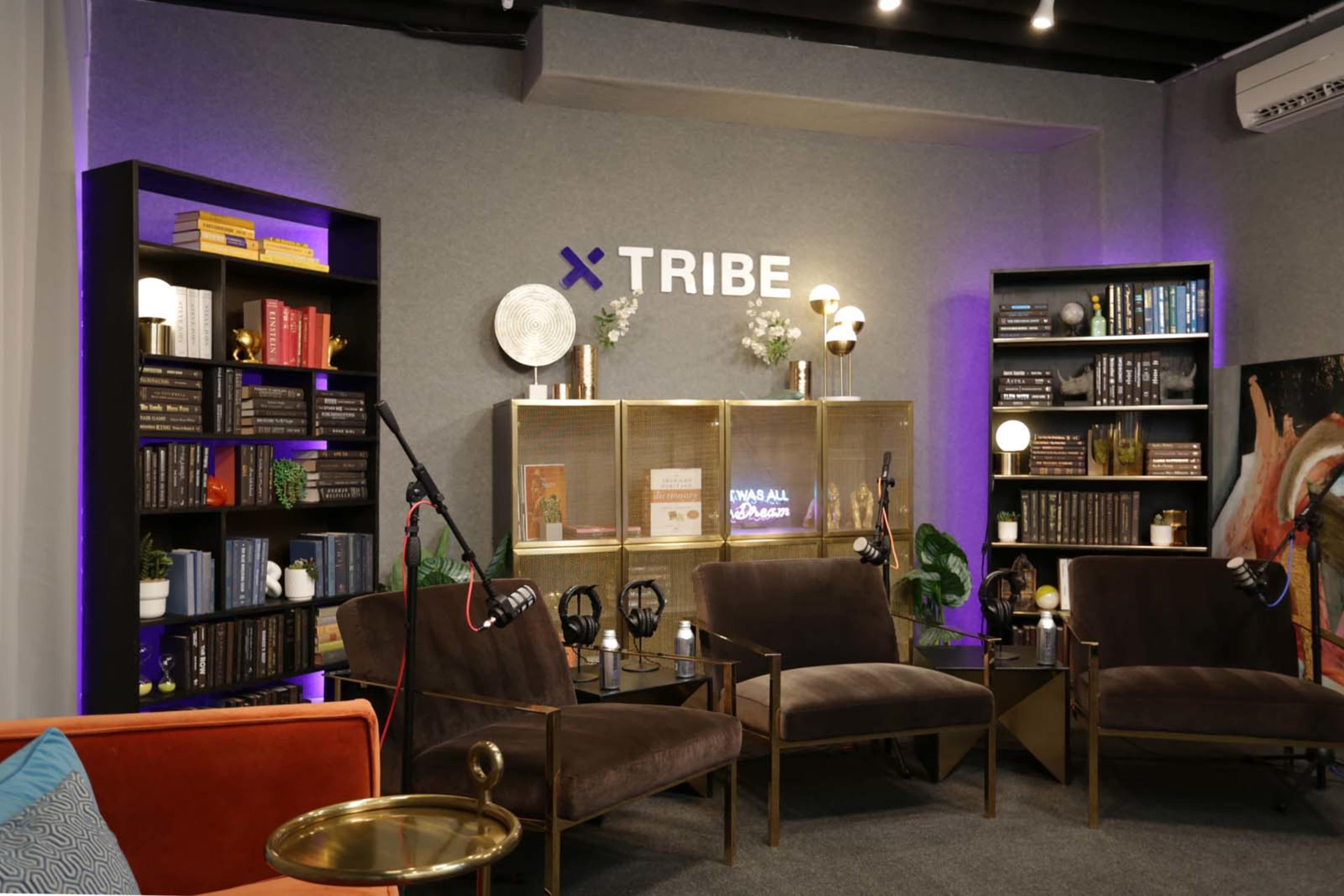 A modern lounge area with brown armchairs, bookshelves filled with books, and ambient lighting, all branded with the word "TRIBE" on the wall.