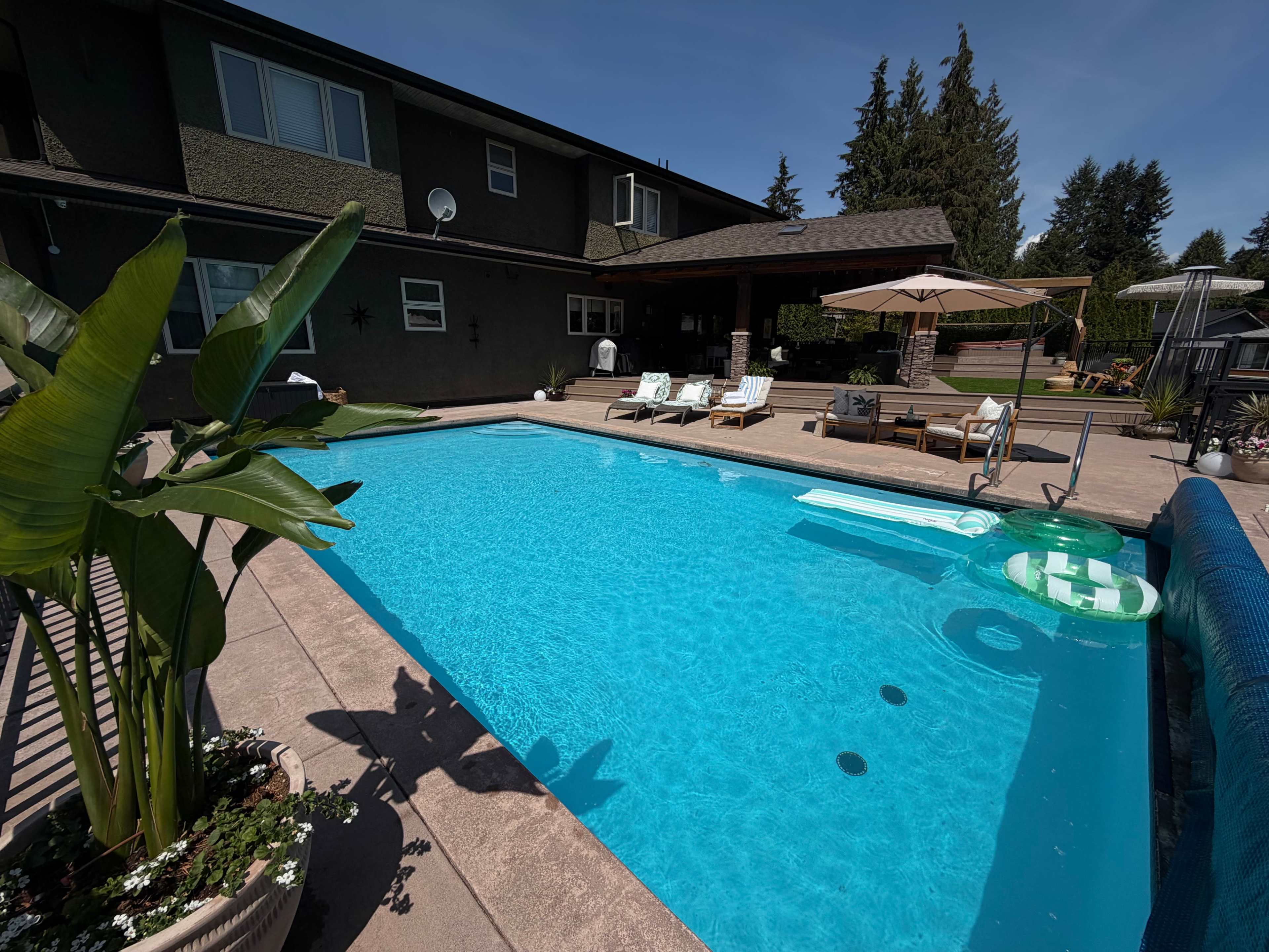 A rectangular swimming pool with clear blue water is surrounded by lounge chairs, a shaded area, and greenery in a residential backyard.