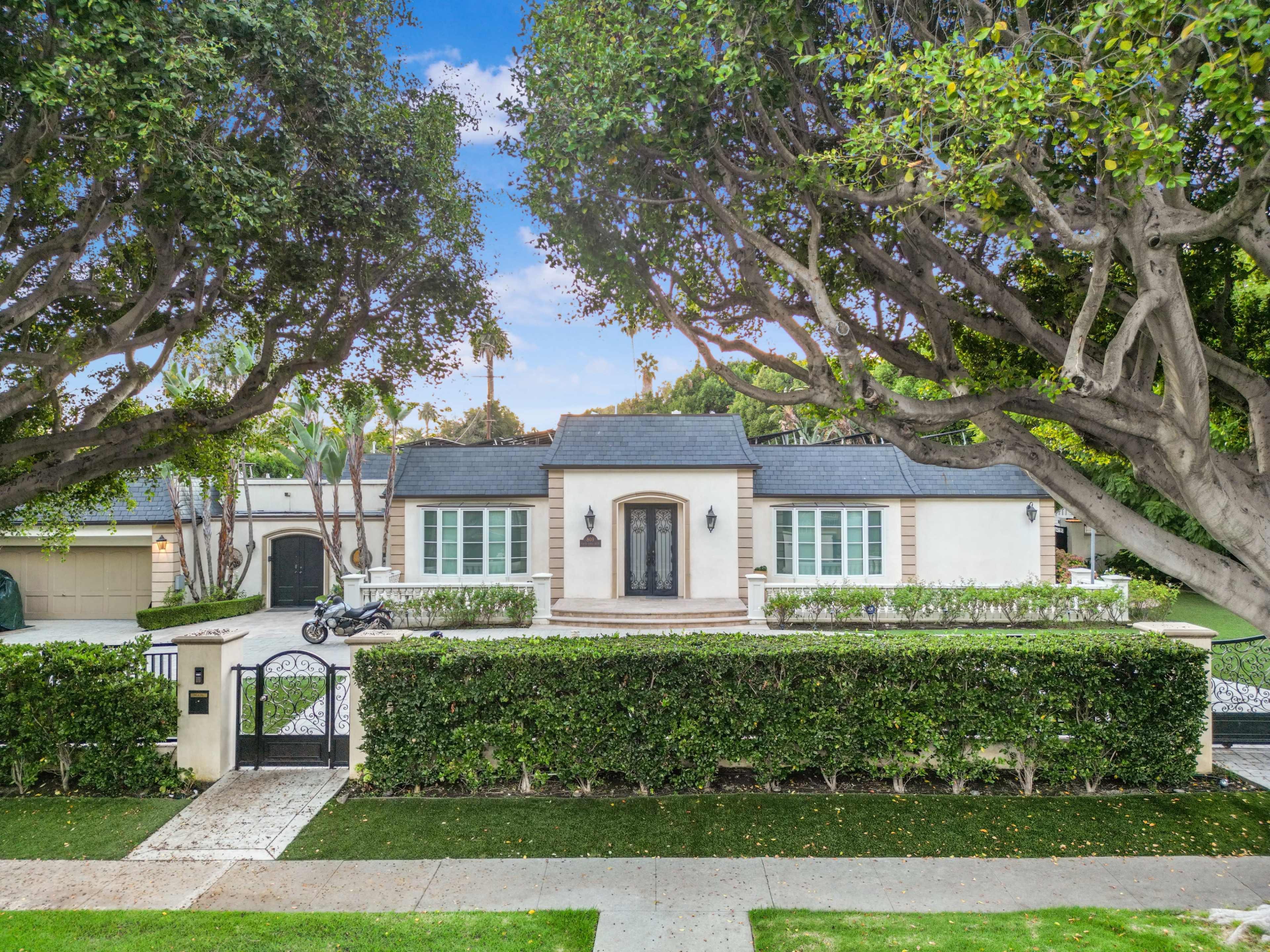 A single-story residential house with a manicured front yard, surrounded by tall trees, features a gated entrance and a driveway on the left.
