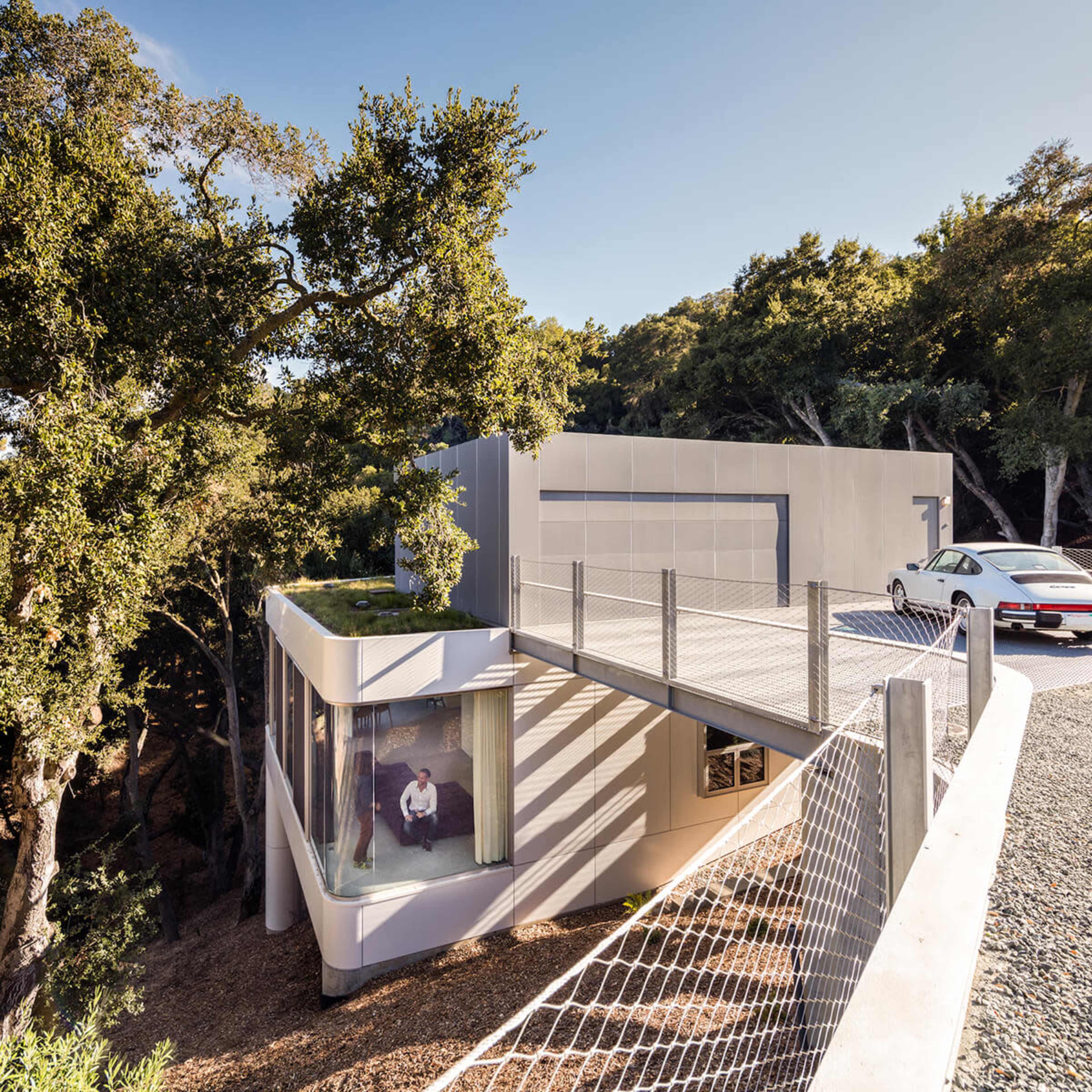 A modern house with a large deck and a glass-walled living area overlooks a wooded area.