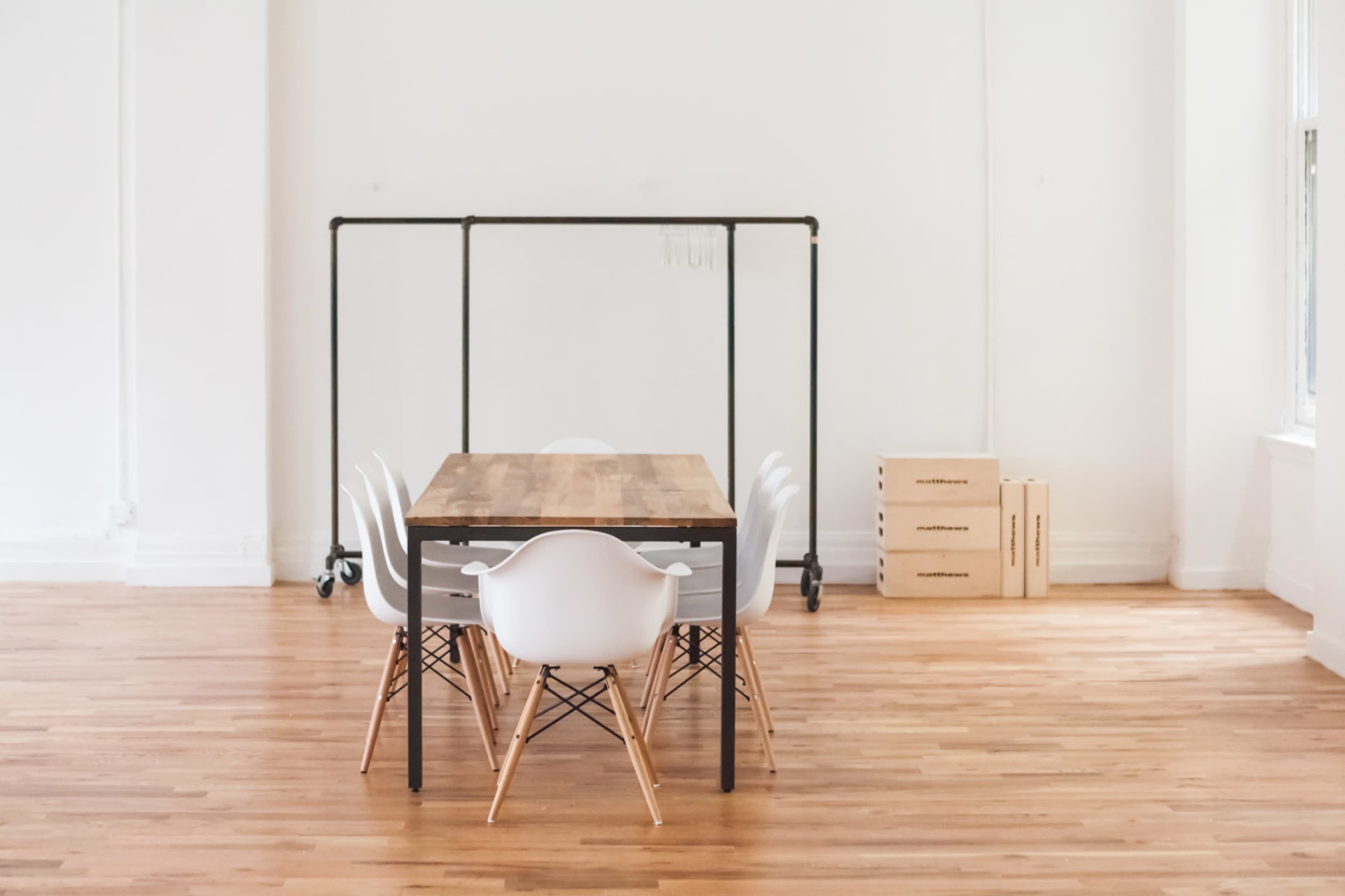 A wooden table surrounded by six white chairs is positioned in a spacious, empty room with light-colored wooden flooring.