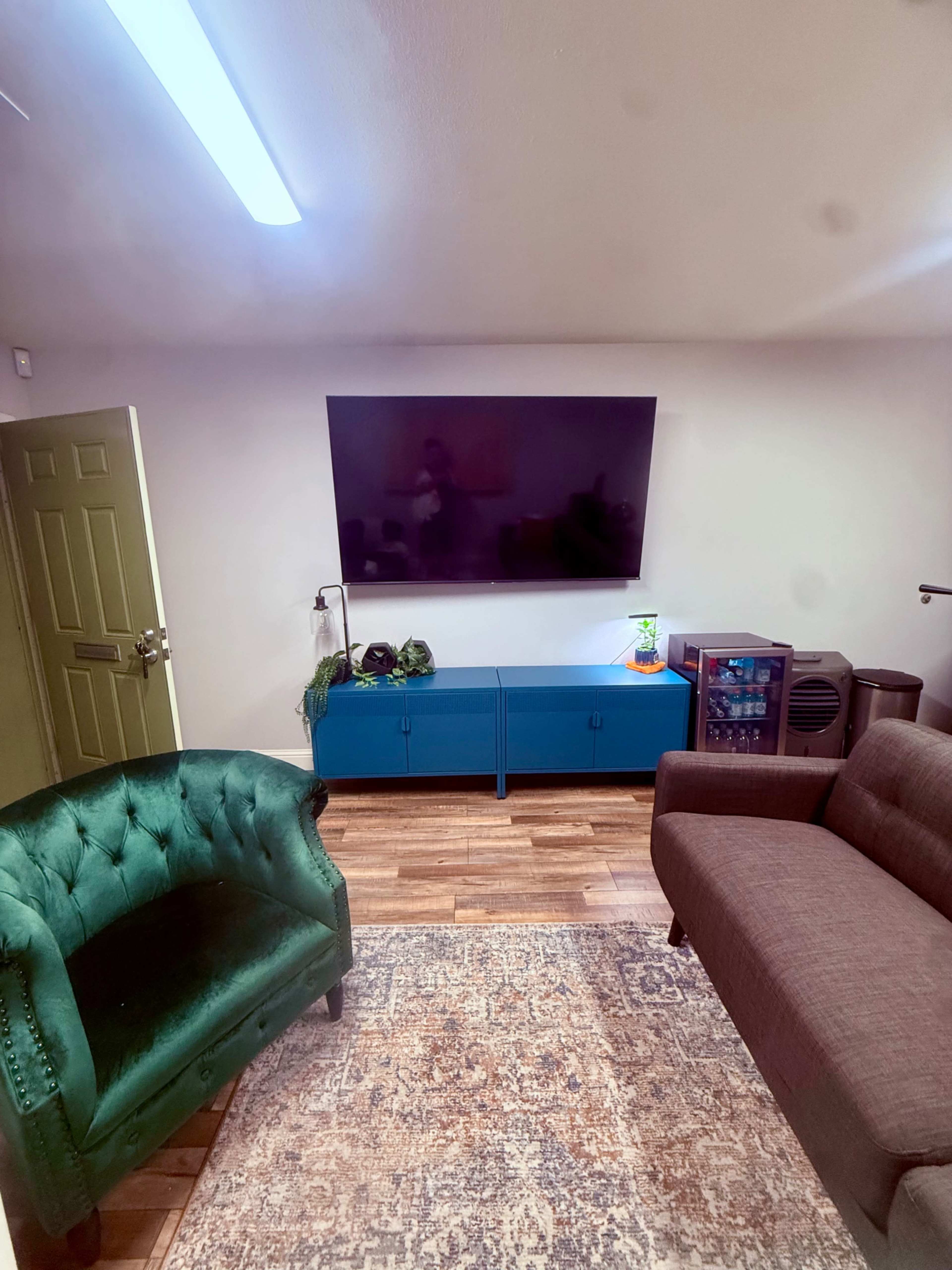 The image shows a living room with a green armchair, a brown sofa, a blue TV stand beneath a large flat-screen television, and wooden flooring.